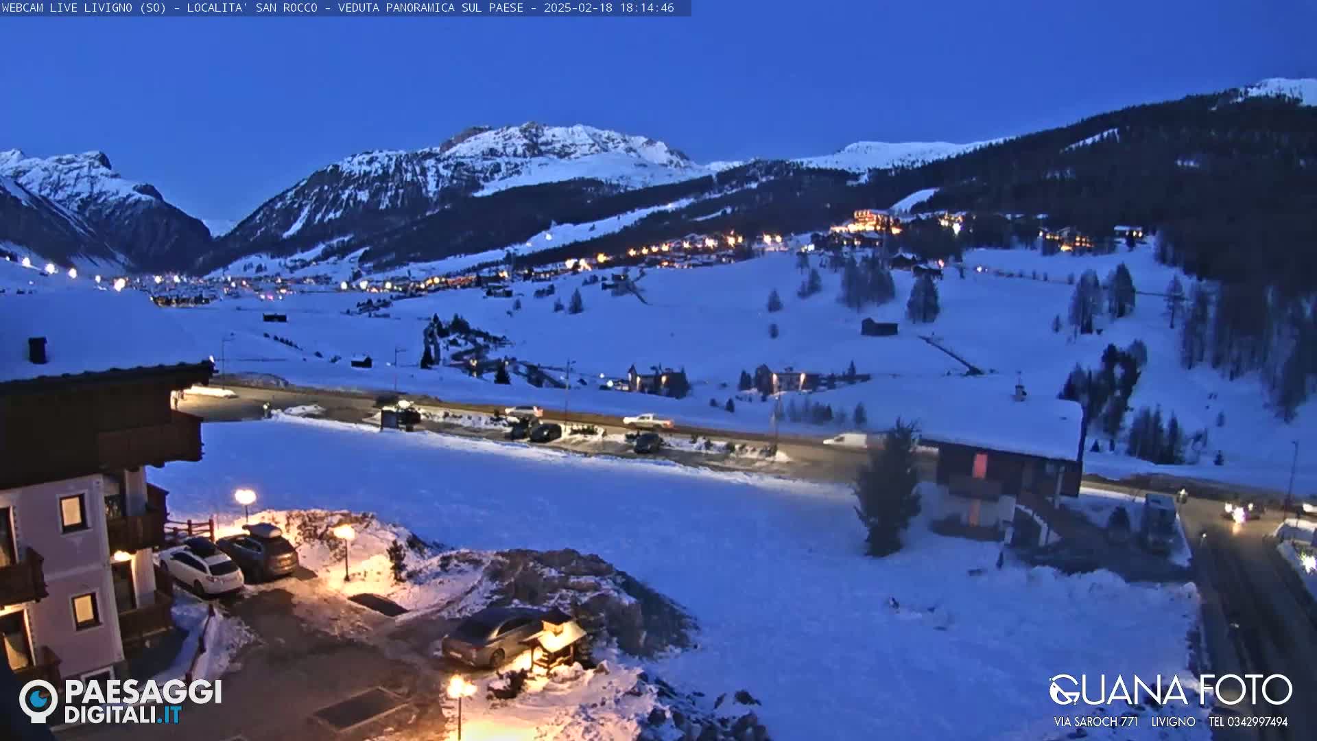 A snow-covered mountain village at twilight, illuminated by scattered lights, with cars parked along a road in the foreground.
