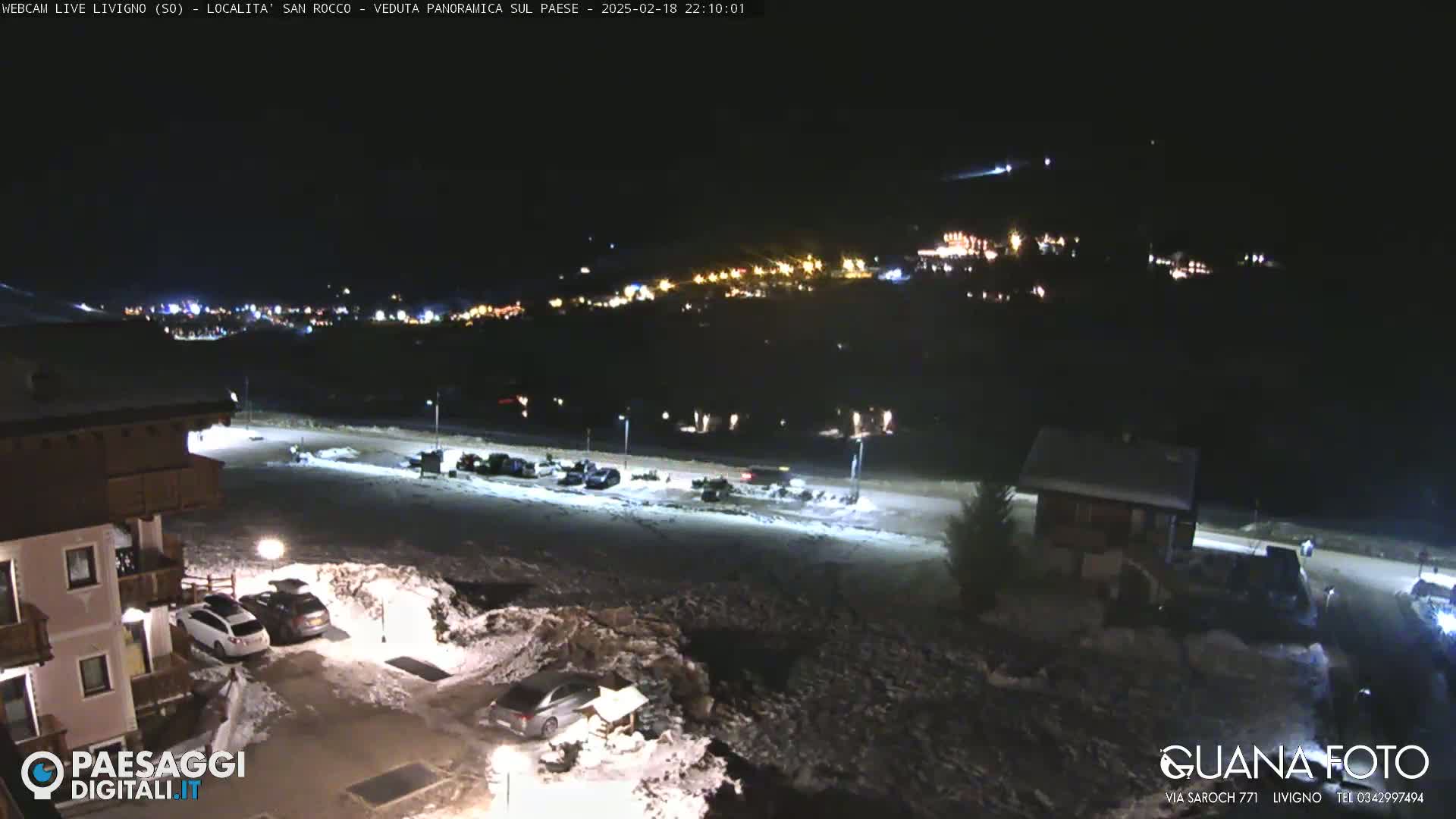 A nighttime, snow-covered scene shows a parking lot with several cars, buildings, and distant lights illuminating a town in the background.