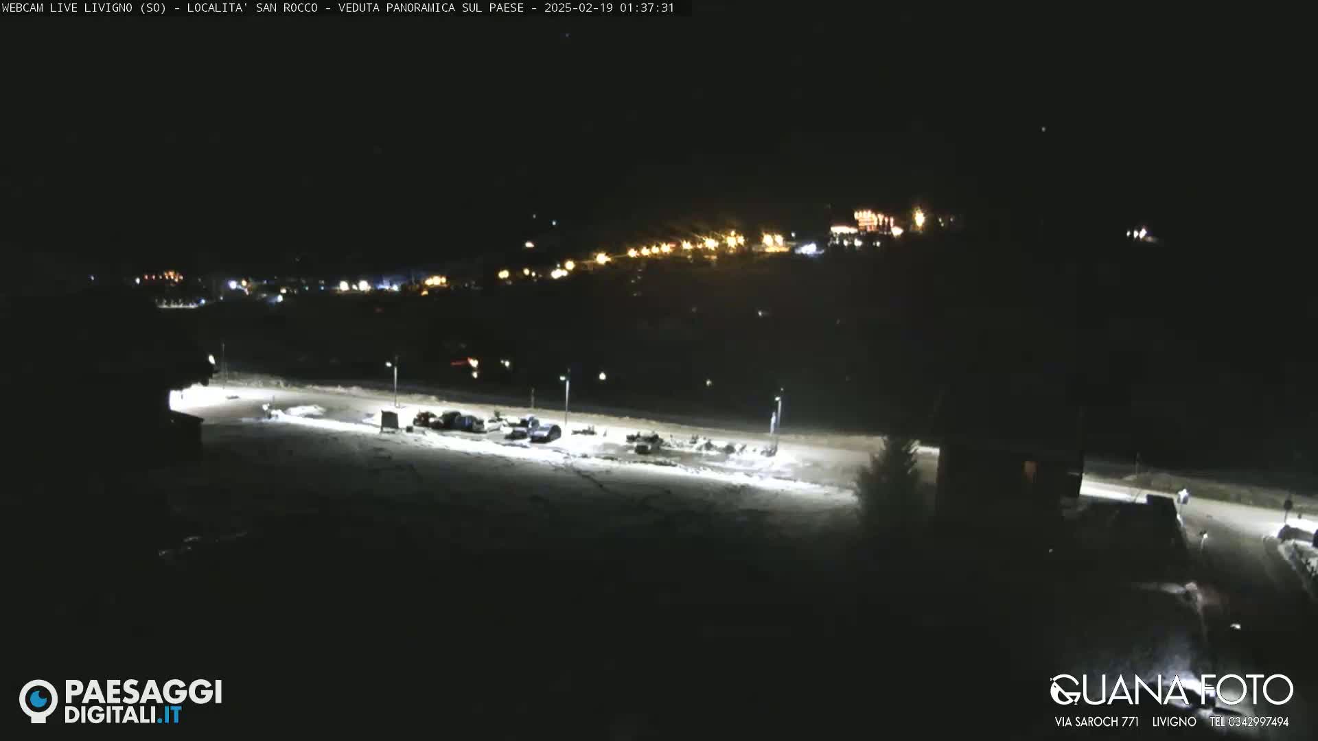 A nighttime, snow-covered parking lot with a few cars is illuminated by artificial lights, overlooking a distant town also lit up at night.