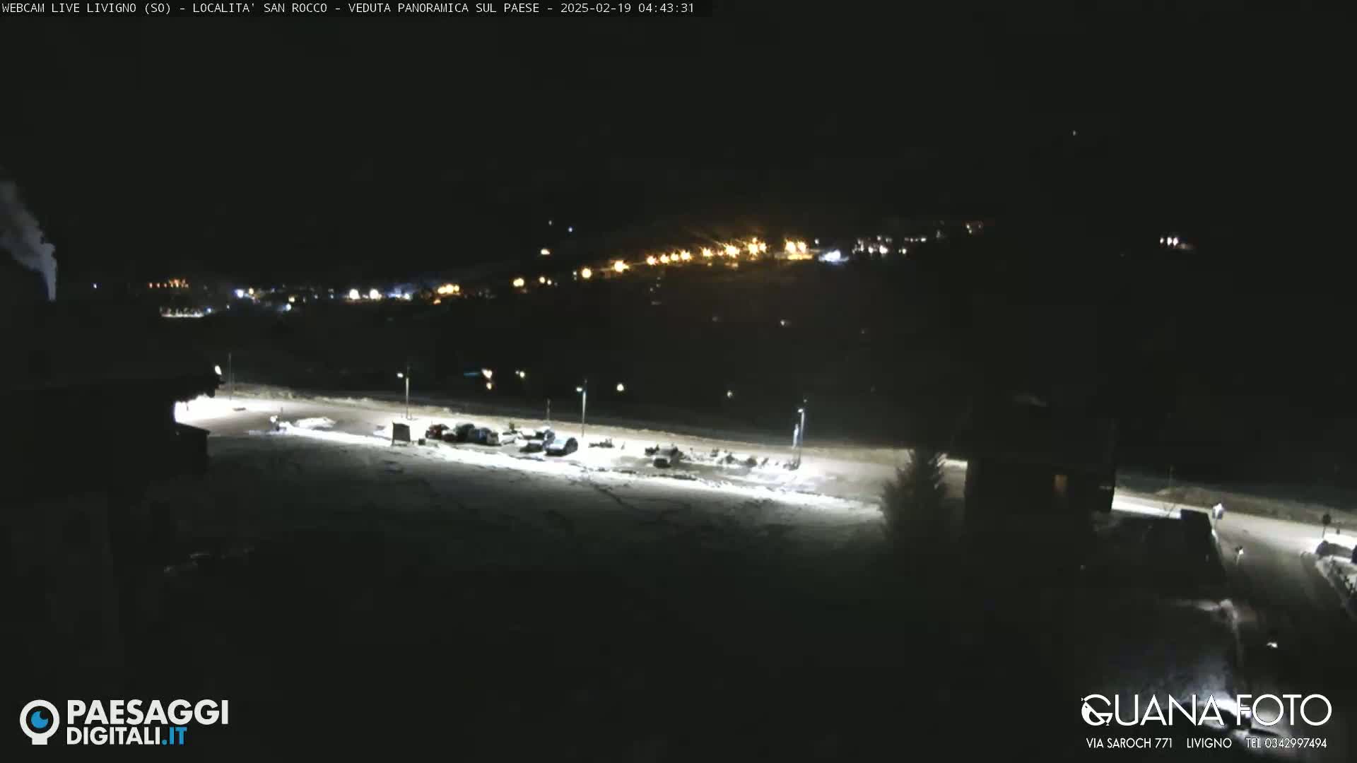 A nighttime, snow-covered parking lot is illuminated by lights, with a distant town faintly visible in the background under a clear, dark sky.