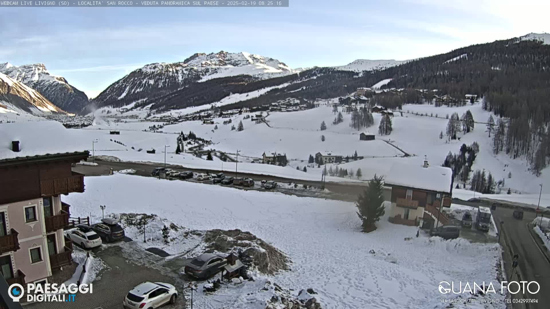 A snow-covered mountain valley town is seen on a partly sunny day, with cars parked in lots and buildings scattered throughout the landscape.