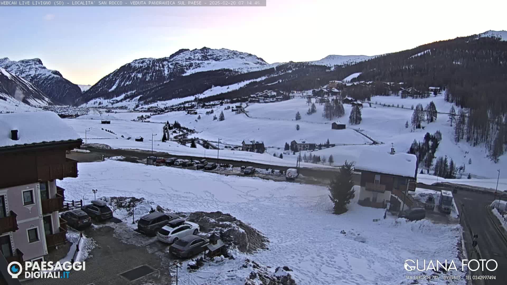 A snow-covered mountain village scene at dawn, with cars parked near buildings and a road winding through the landscape.