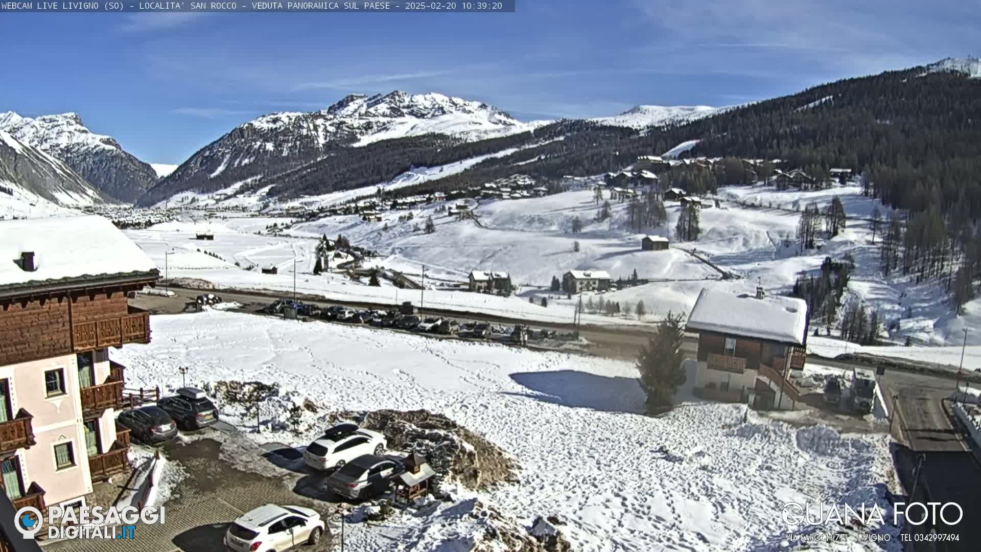 A snow-covered valley town is viewed from a hillside, under a clear blue sky, with several buildings, cars parked in a lot, and mountains in the background.
