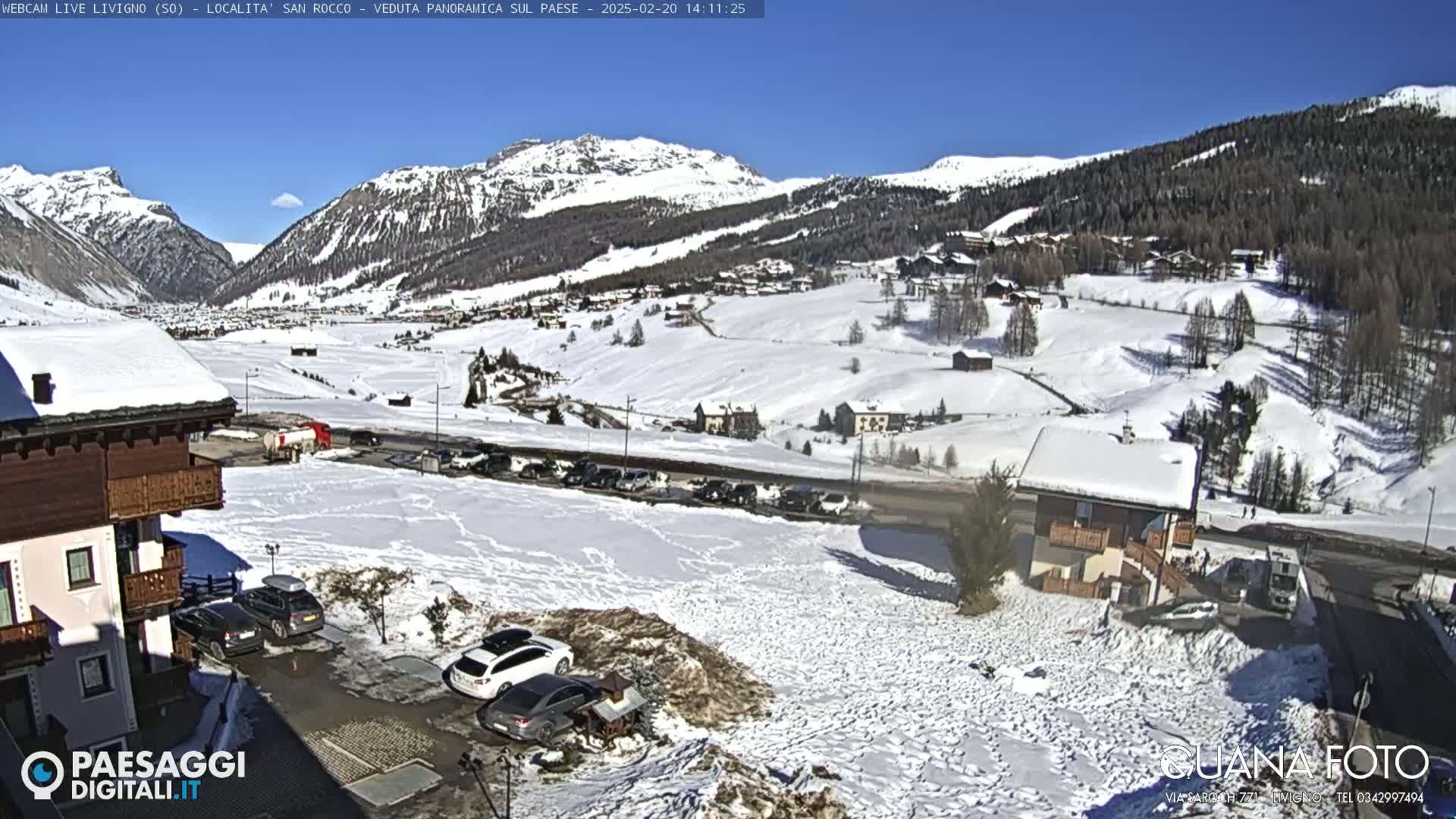 A snow-covered mountain village is viewed from a hillside on a sunny day, with cars parked in a lot and buildings in the foreground.