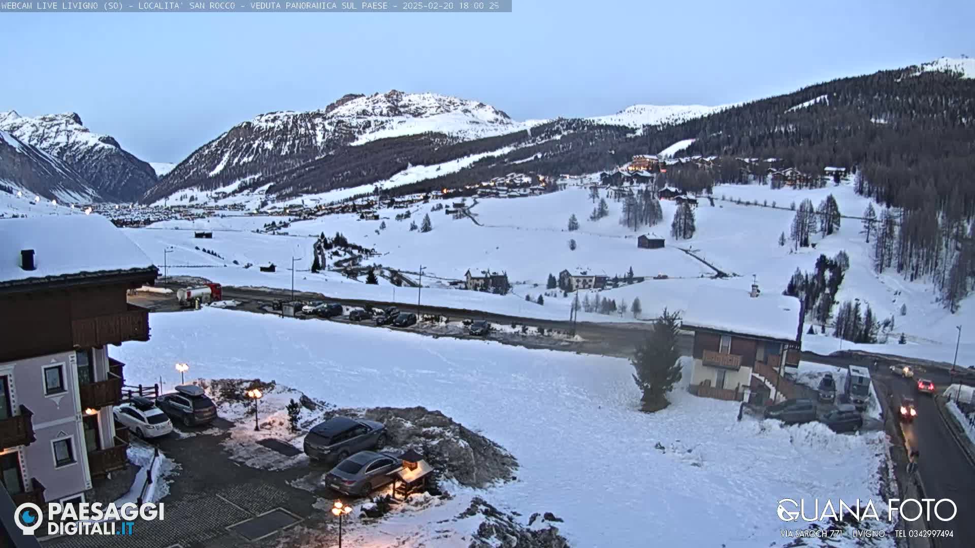 A snow-covered mountain village is seen at twilight, with cars parked near a building in the foreground.