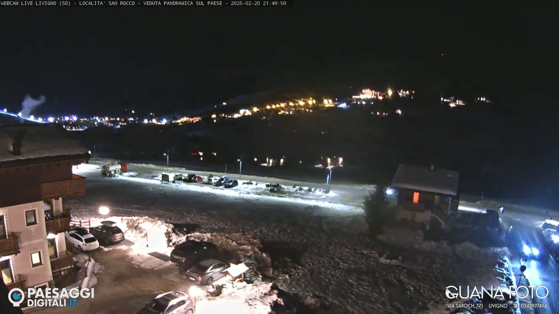 A nighttime, snow-covered parking lot and surrounding buildings are illuminated, with a distant town visible on a hillside under a clear, dark sky.