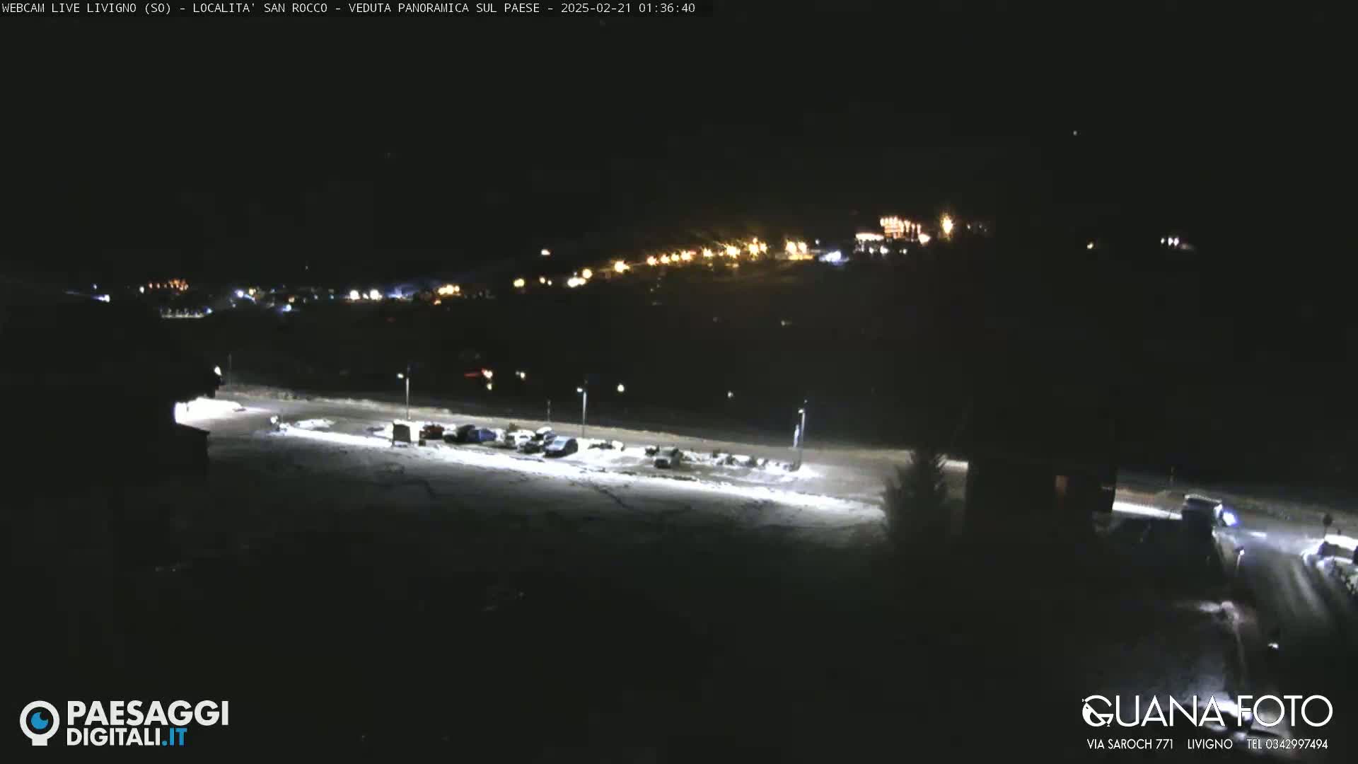 A nighttime, snow-covered parking lot and road are illuminated by streetlights, with a hillside town faintly visible in the distance under a dark sky.