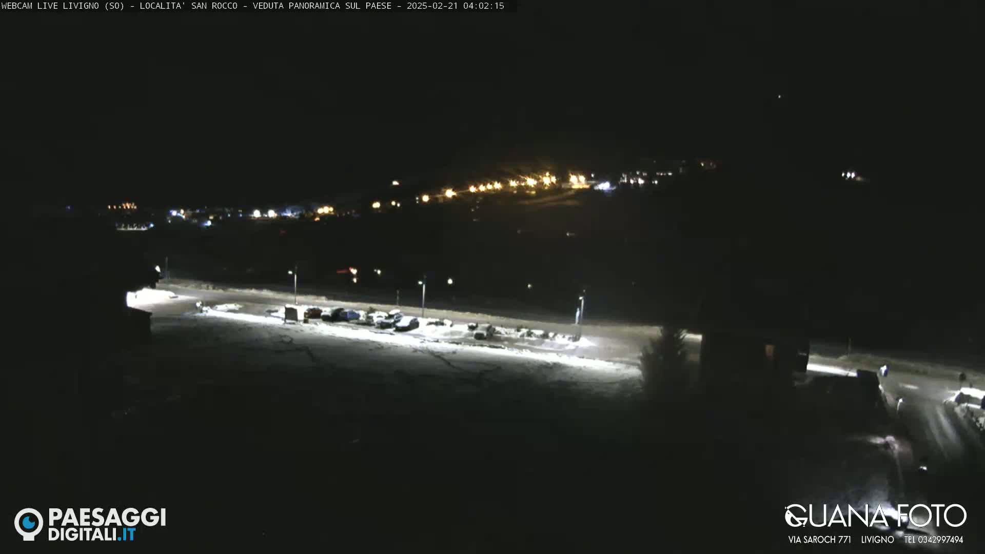A snow-covered parking lot with several cars is illuminated at night, with a brightly lit town visible in the distance under a clear, dark sky.