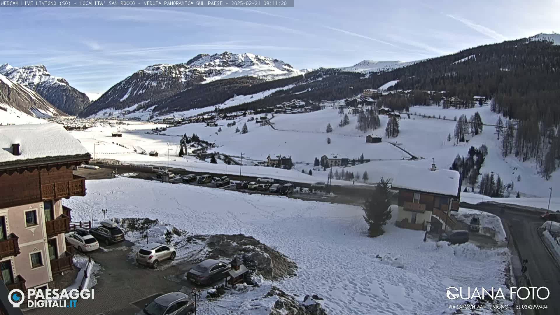 A snow-covered mountain village is seen from a high vantage point on a clear, sunny day, with several cars parked near a building in the foreground.