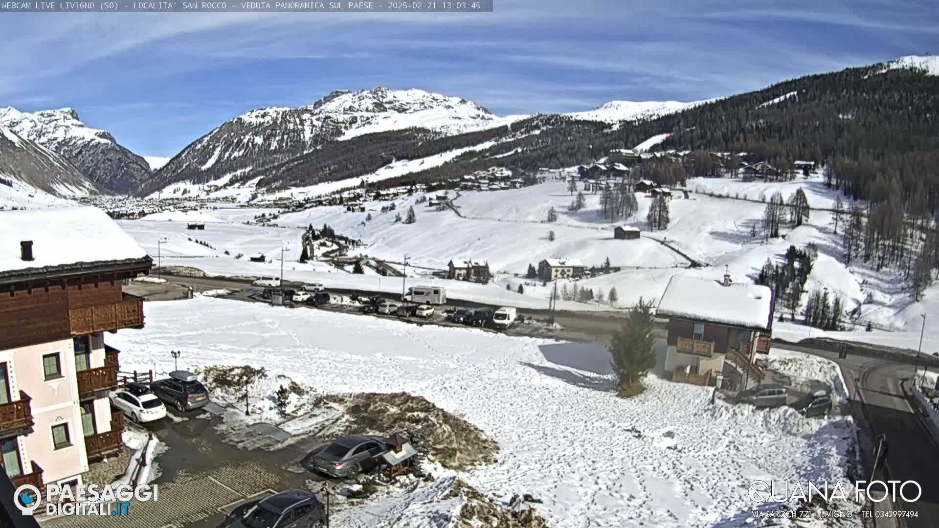 A snow-covered mountain village is viewed from a slightly elevated position on a sunny day, with cars parked in a lot near several buildings.