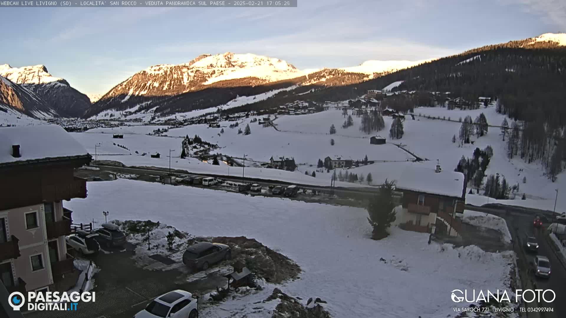 A snow-covered mountain valley at dusk, with a few buildings and cars visible, and a building in the foreground.
