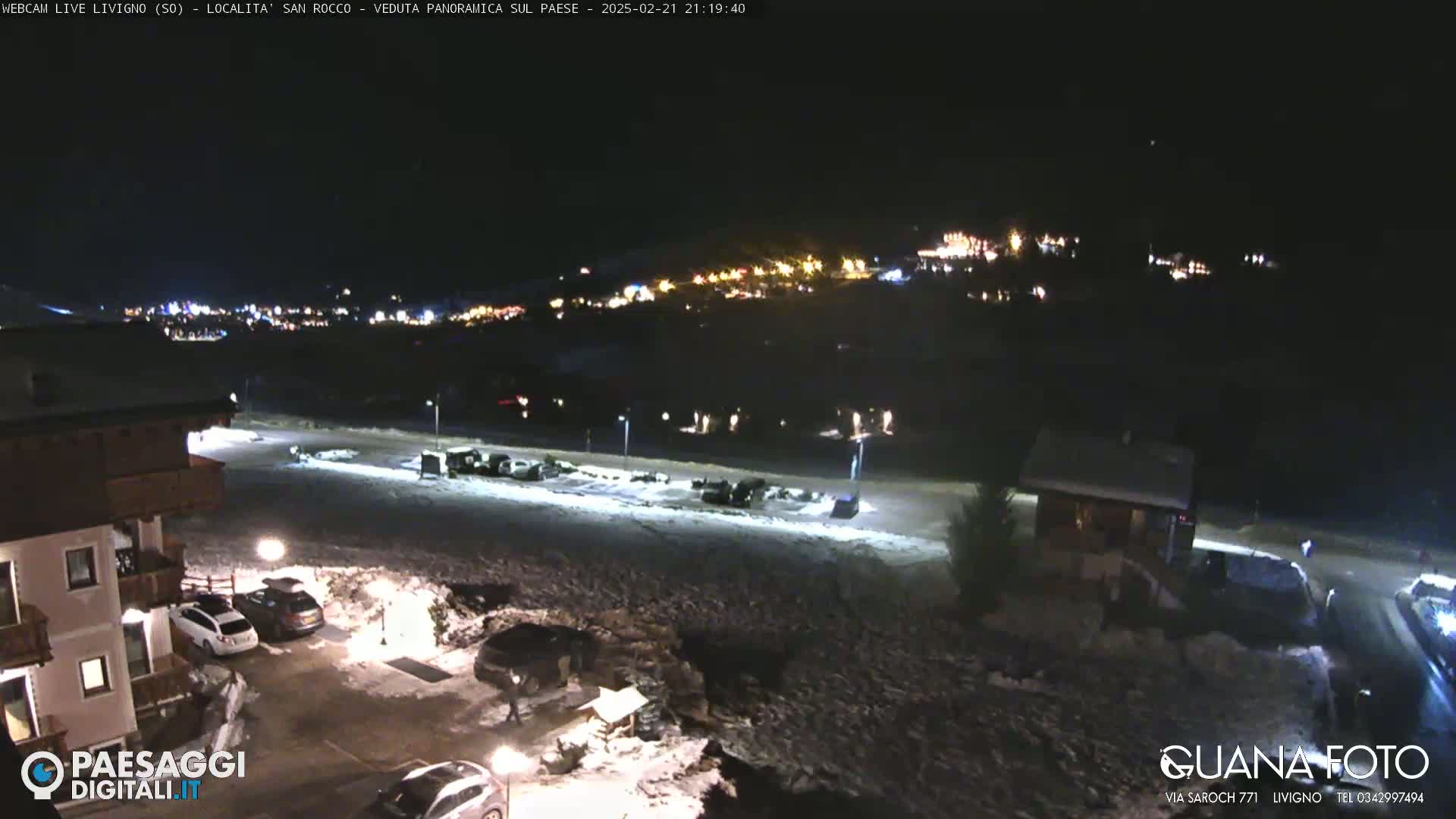 A nighttime view of a snow-covered parking lot and surrounding buildings, with a village illuminated in the distance under a clear, dark sky.