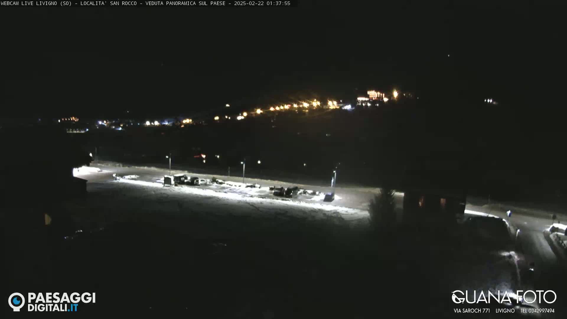 A snow-covered parking lot is illuminated at night, with a distant village glowing faintly in the background.
