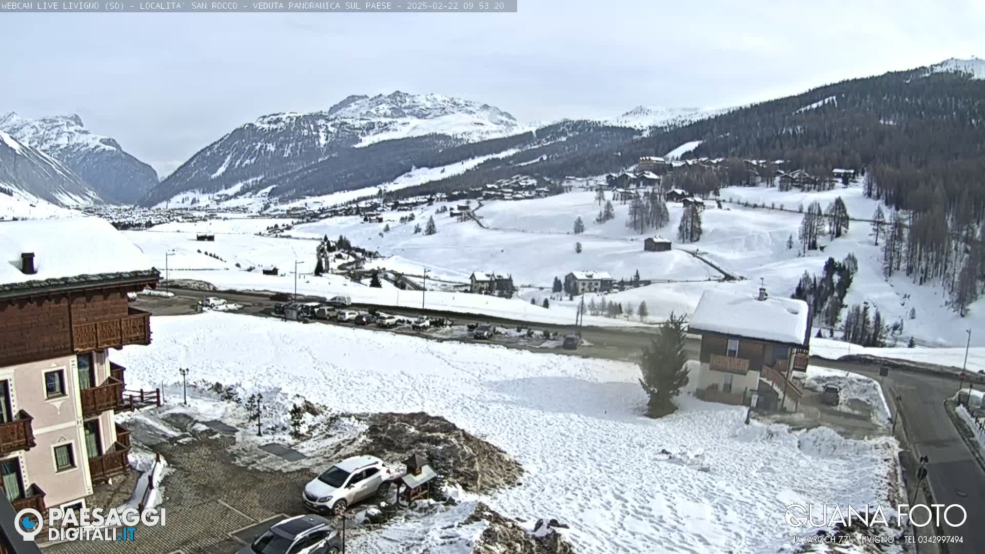 A snow-covered valley town is viewed from a slightly elevated position, showing buildings, roads, and cars under a cloudy sky.