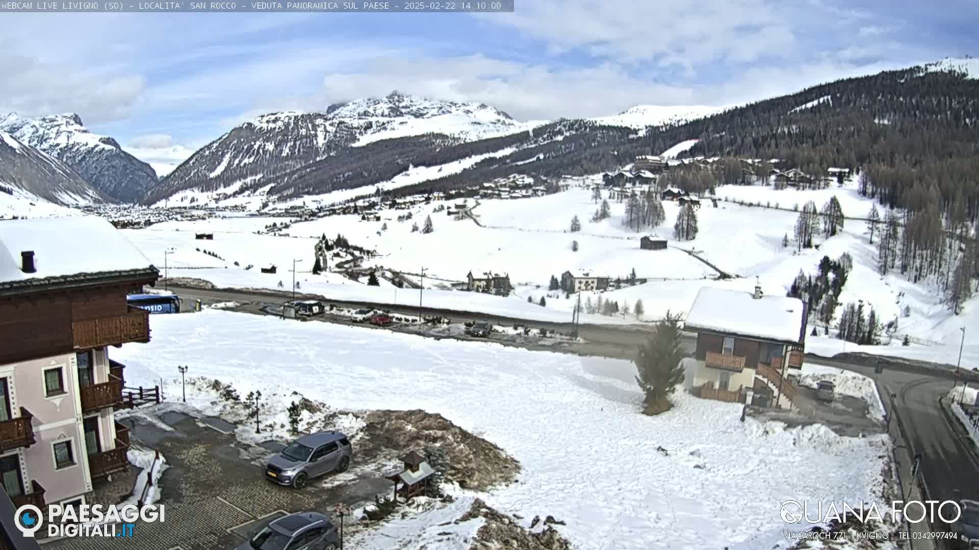 A snow-covered mountain village is viewed from a slightly elevated position, with cars parked in a nearby lot under a partly cloudy sky.