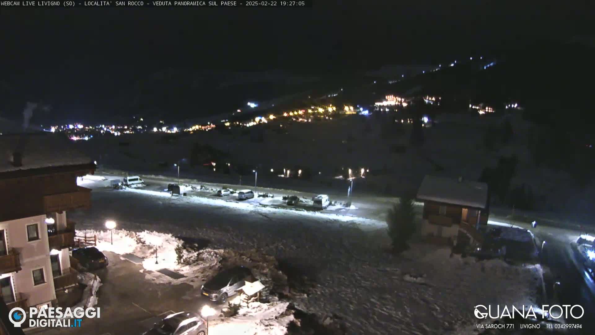 A nighttime, snow-covered parking lot and village are illuminated, with a hill showing distant lights in the background.