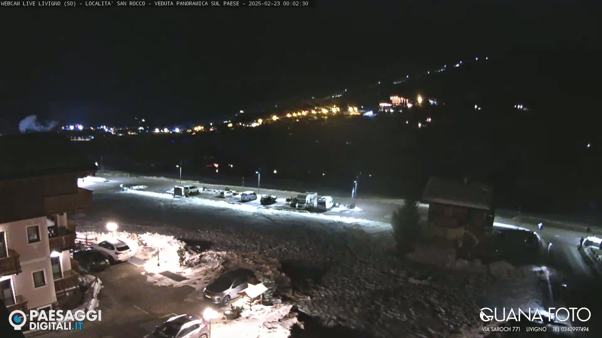 A nighttime, snow-covered parking lot and surrounding buildings are illuminated, with a hillside of lights visible in the distance.