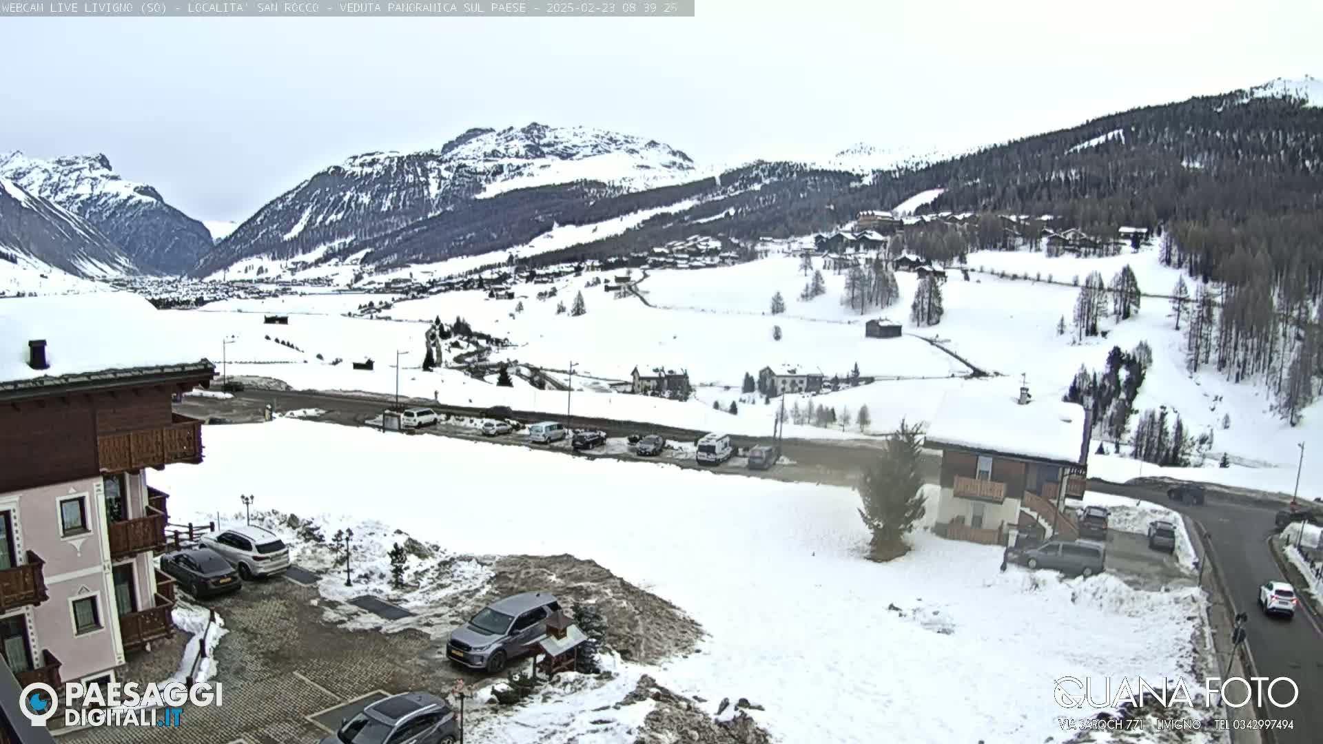 A snow-covered mountain valley scene with several buildings, cars in parking lots, and a partly cloudy sky.