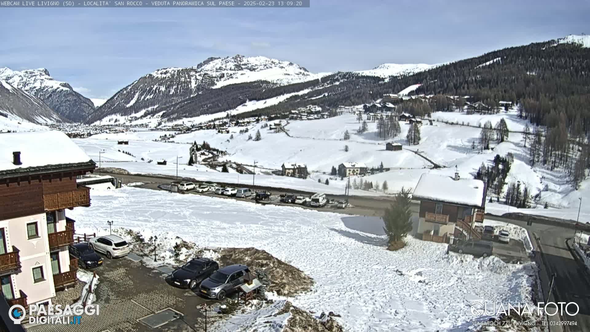 A snow-covered mountain village is viewed from a hillside, with cars parked in a lot below, under a clear blue sky.