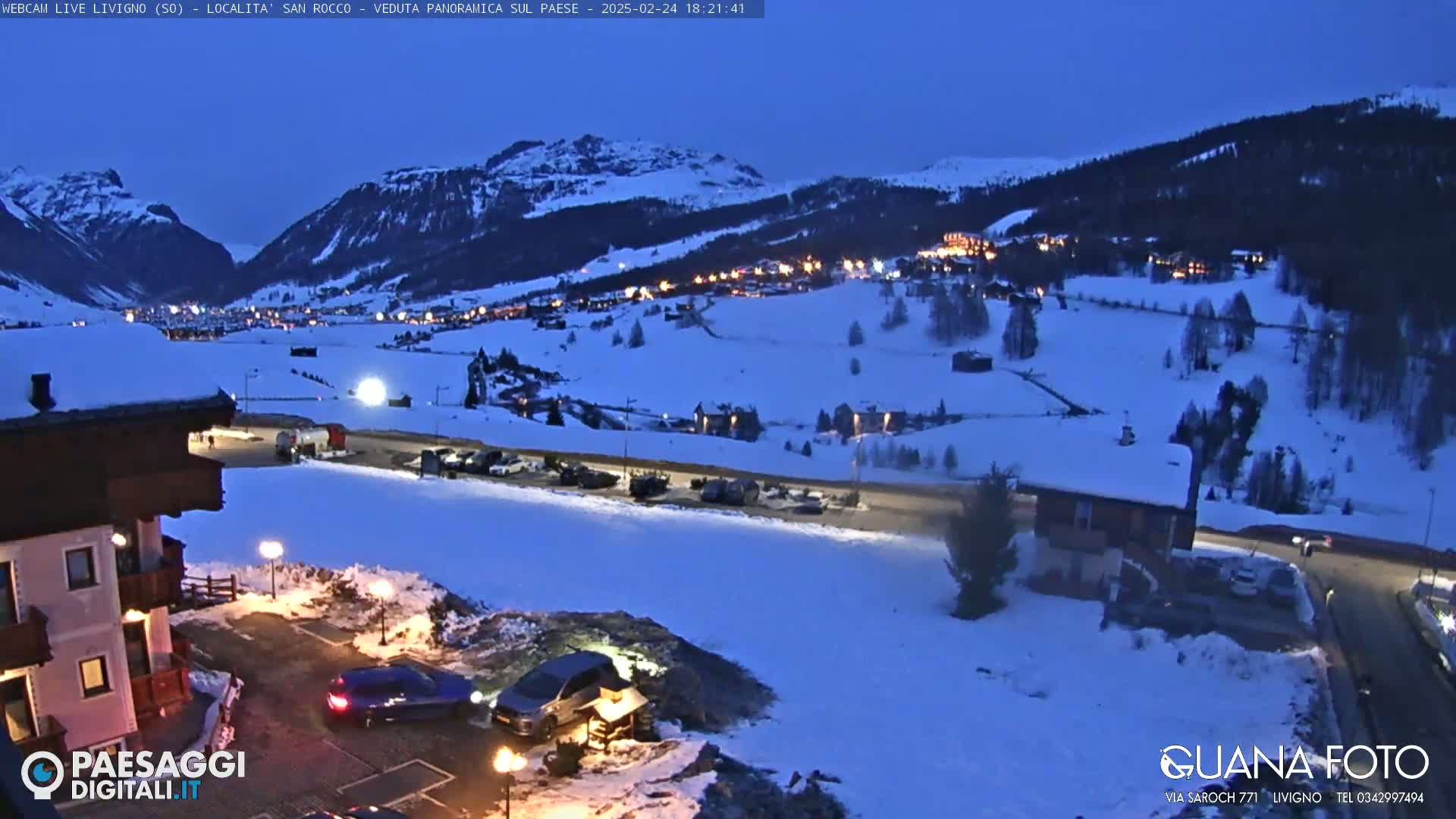 A snow-covered village at twilight, nestled in a valley between mountains, with cars parked in lots nearby.