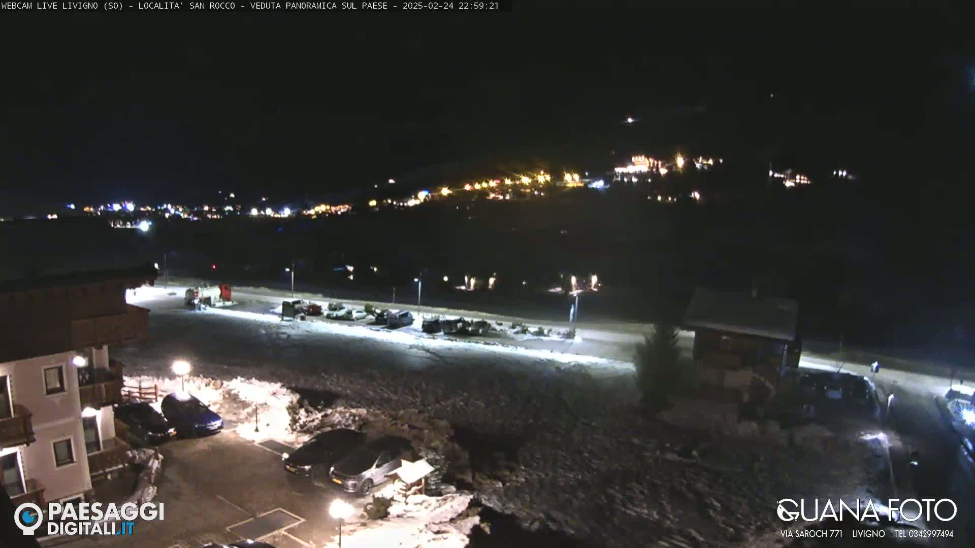 A nighttime, snow-covered parking lot and village are illuminated by streetlights and building lights, with a hilltop town visible in the distance.