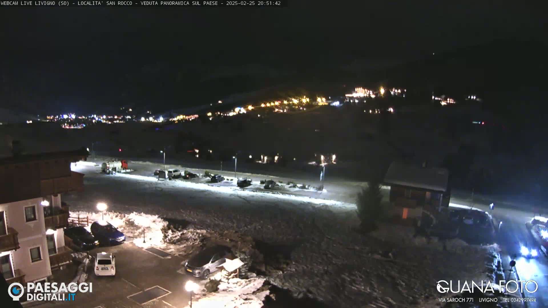 A nighttime, snowy scene shows a village in the distance illuminated by lights, with cars parked in a lot in the foreground and a building nearby.