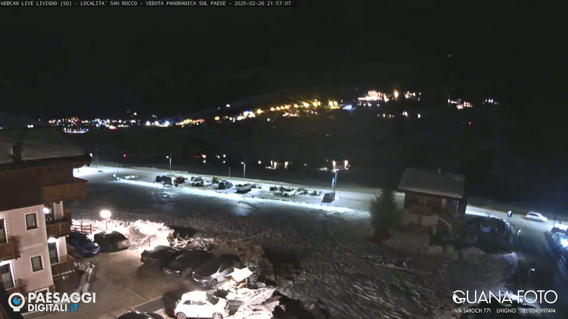 A nighttime view of a snow-covered parking lot and town, illuminated by streetlights and distant building lights.