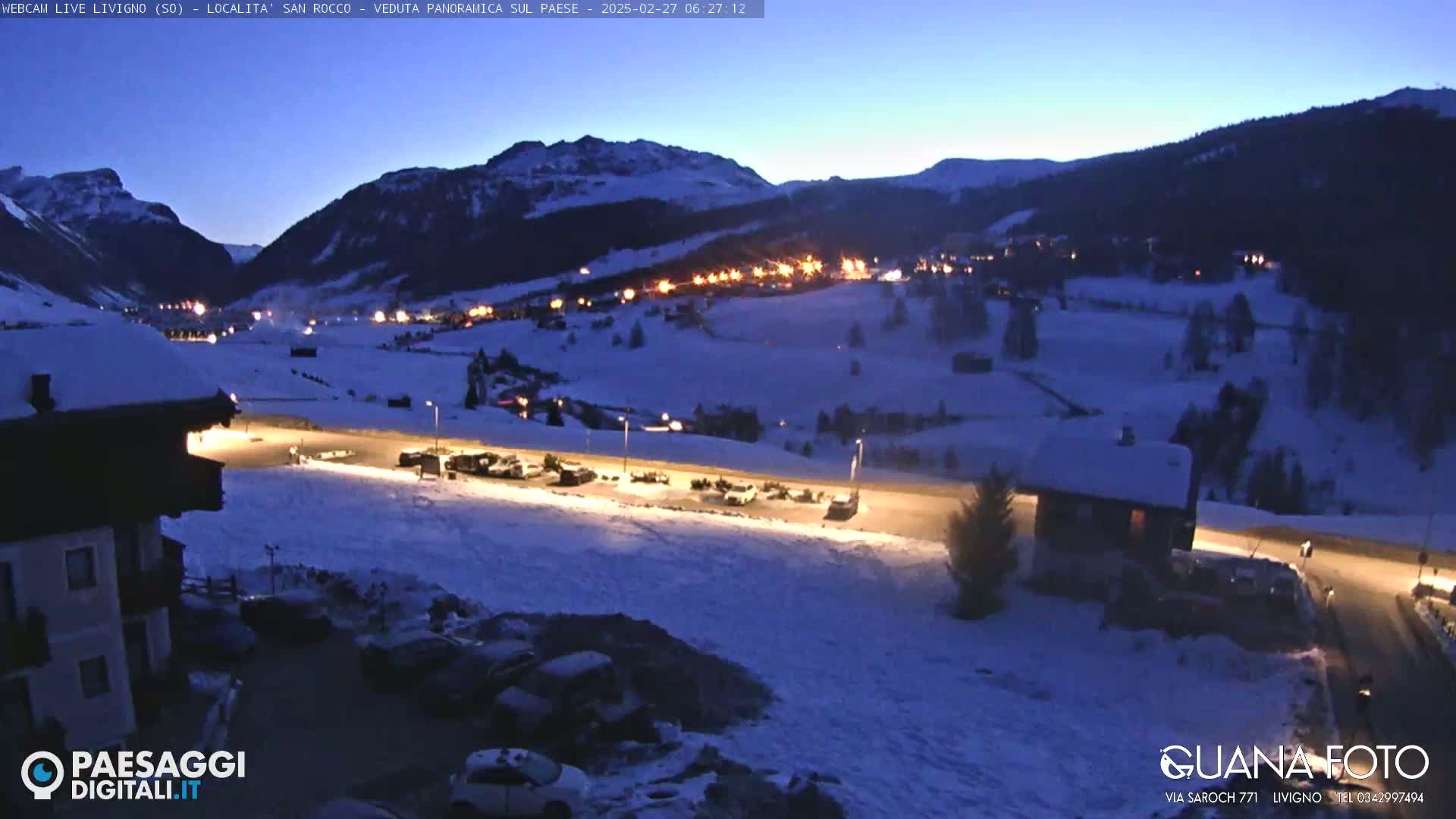 A snow-covered mountain village at twilight, illuminated by artificial lights, with cars parked in a lot in the foreground.