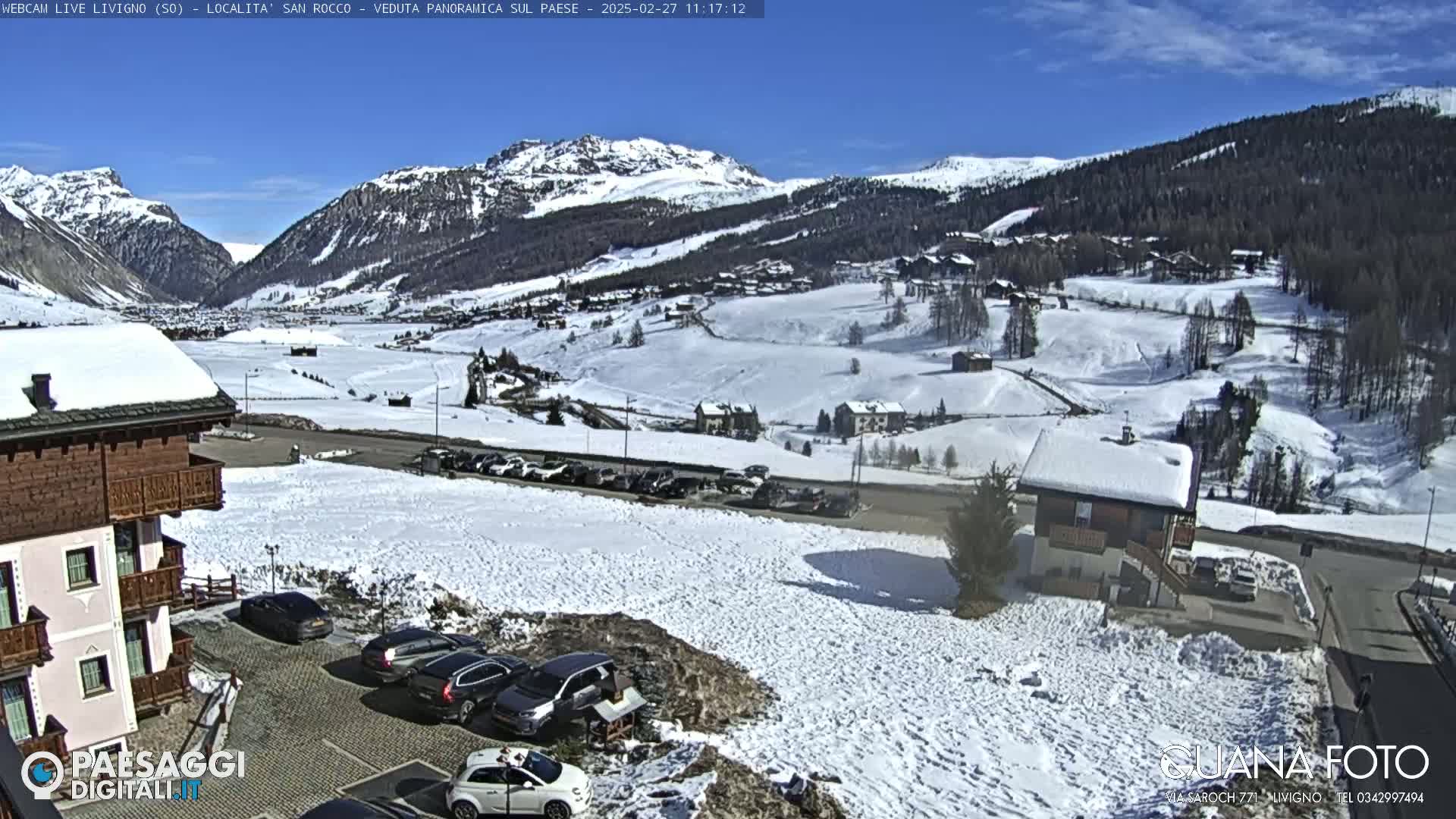 A snow-covered mountain valley town is viewed from a slight elevation on a sunny day, with cars parked in a lot near a building in the foreground.