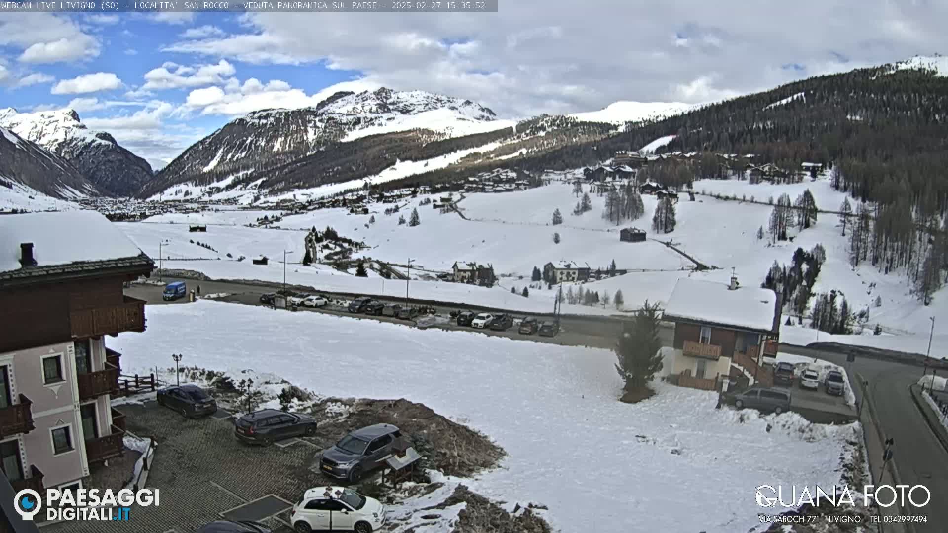 A snowy mountain village is viewed from a vantage point, with partly cloudy skies and several cars parked in a lot in the foreground.