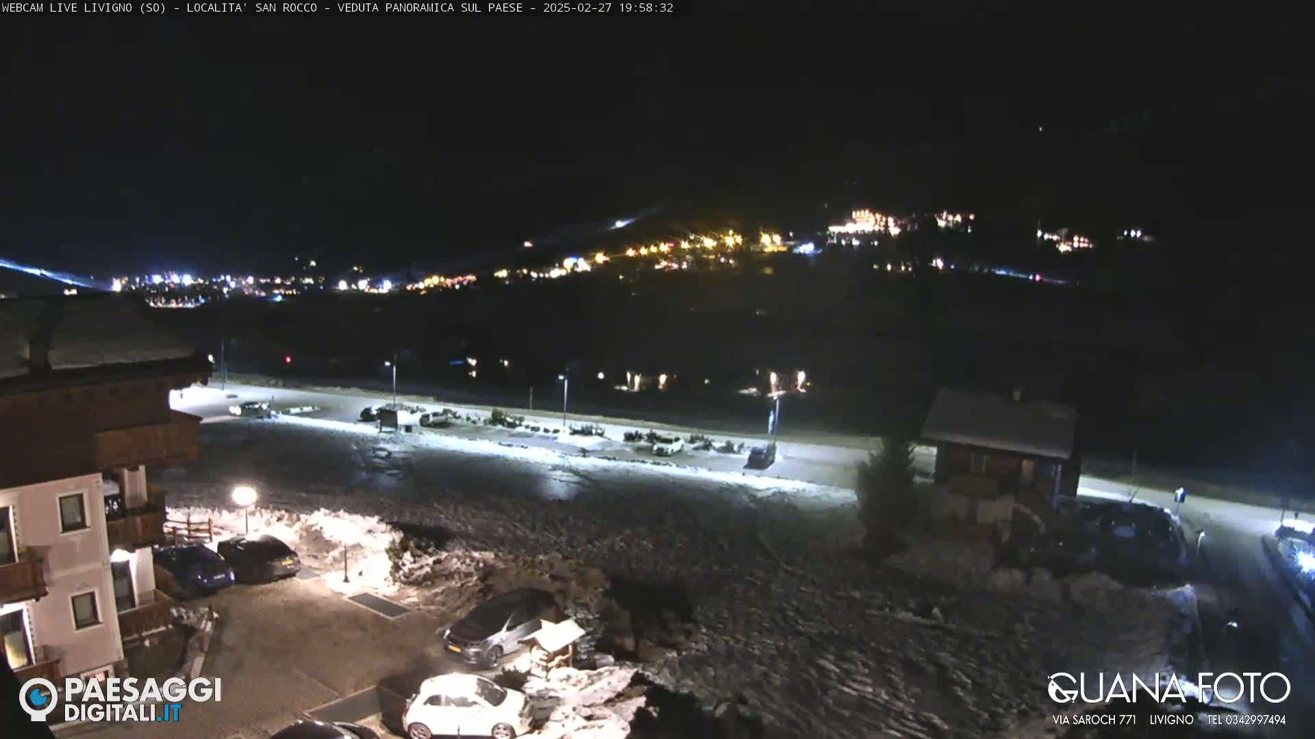 A nighttime view of a snow-covered parking lot and buildings, with a town illuminated in the distance under a clear, dark sky.