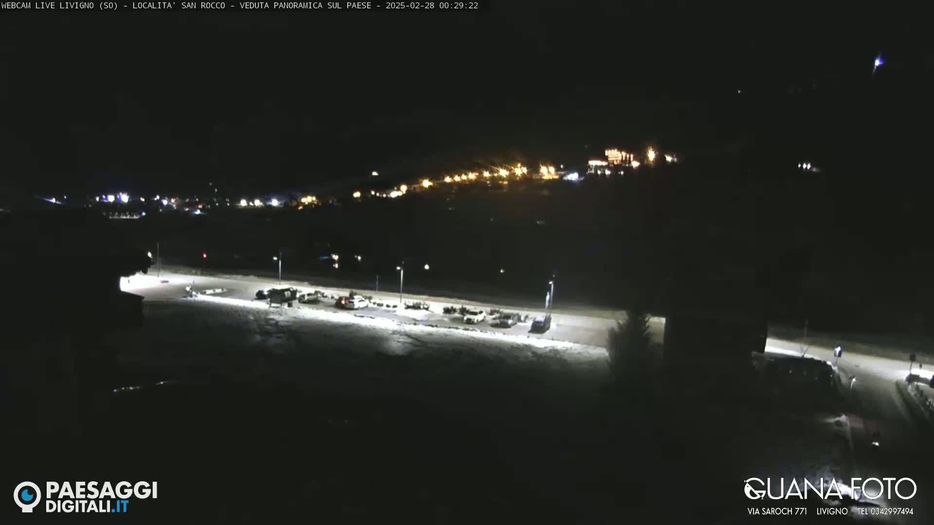 A nighttime, snow-covered parking lot with a few cars, illuminated by streetlights, overlooks a distant town with lights visible in the background.