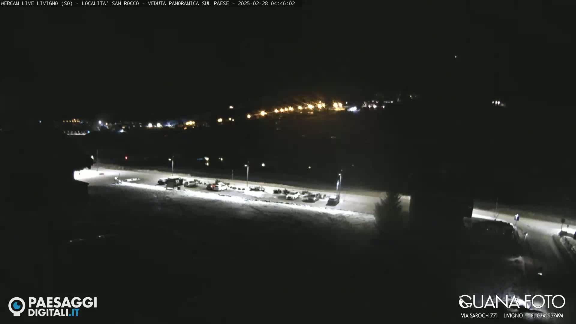 A nighttime, snow-covered parking lot with several cars is visible, and in the distance, a town's lights illuminate the hillside.