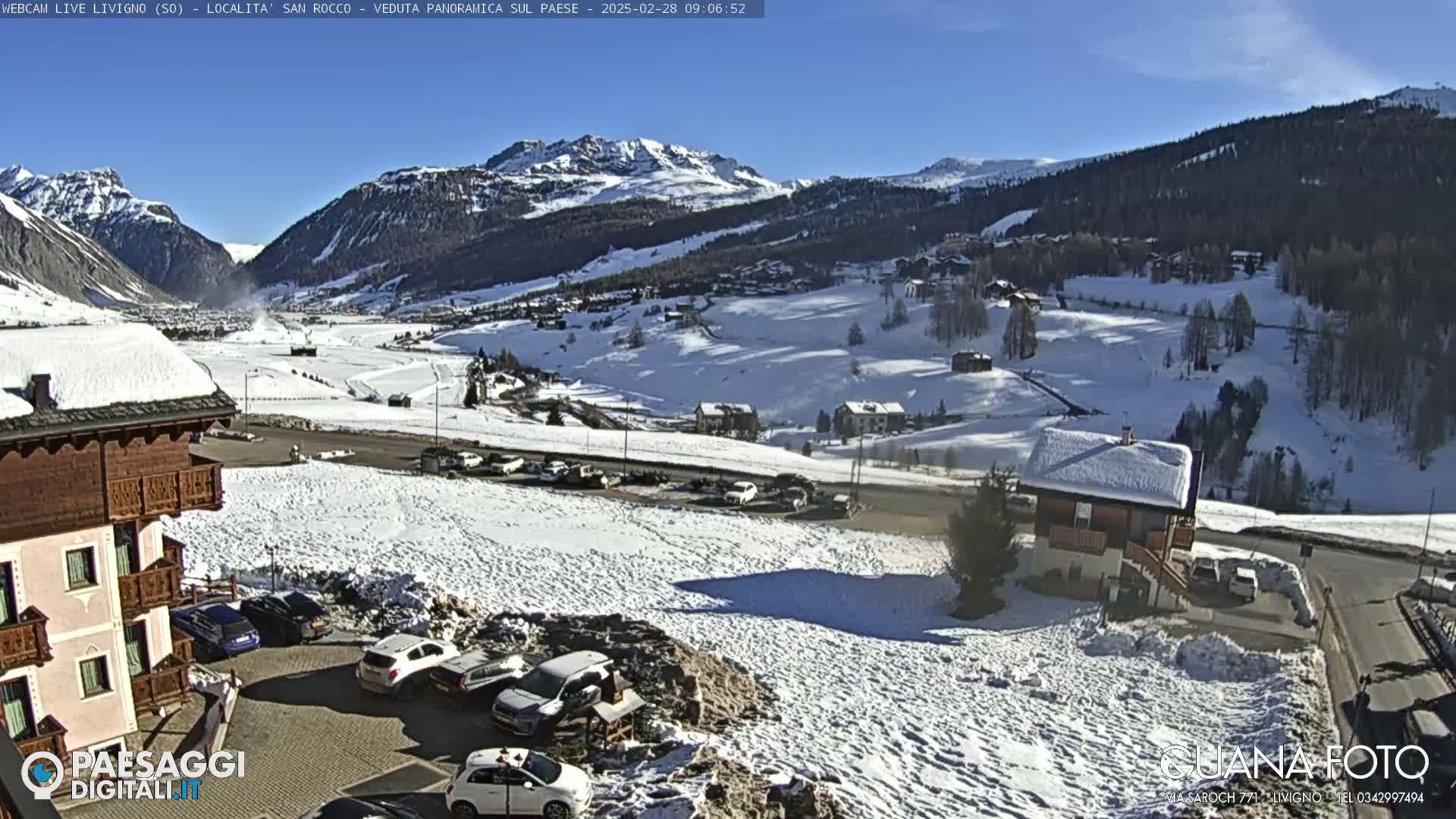 A snow-covered mountain valley on a sunny day, with cars parked in a lot near a building and other buildings visible in the distance.