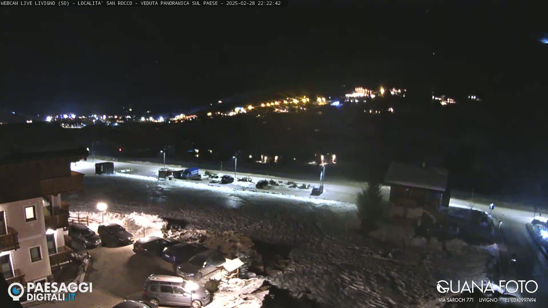 A nighttime, snow-covered parking lot and a town illuminated by lights in the distance.