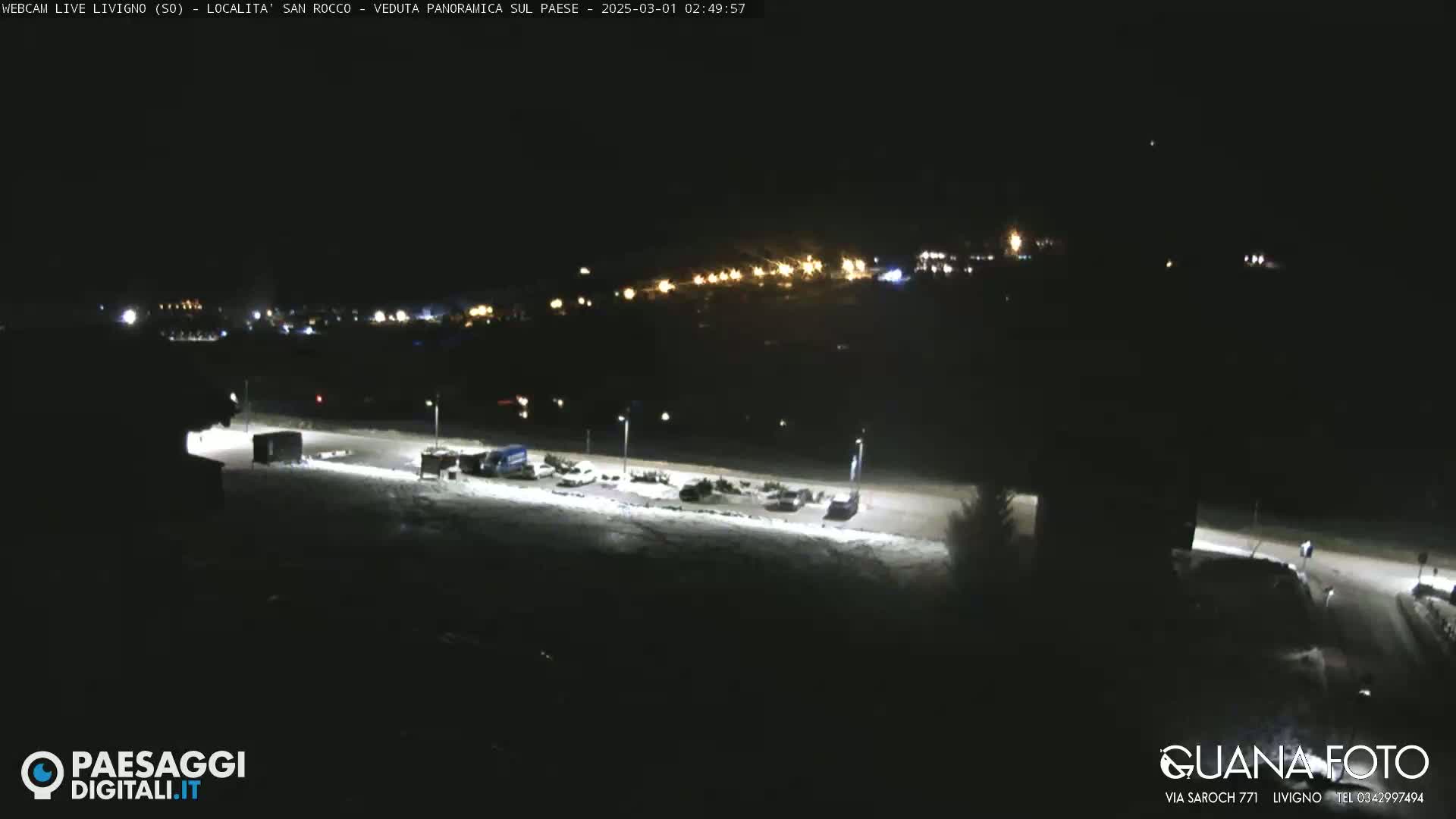 A nighttime, snow-covered parking lot and road are illuminated by streetlights, with a distant village faintly visible in the background under a clear sky.