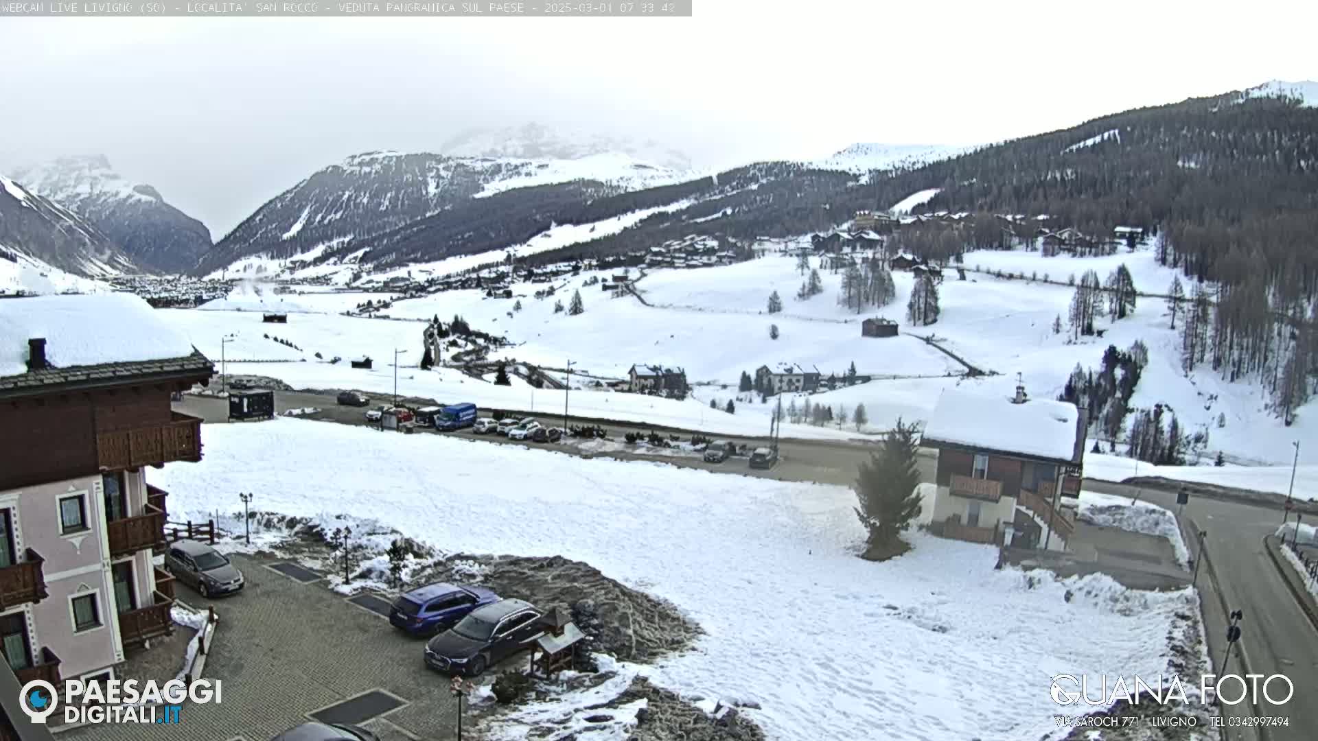 A snow-covered mountain village scene with cars parked in a lot near a building, under an overcast sky.