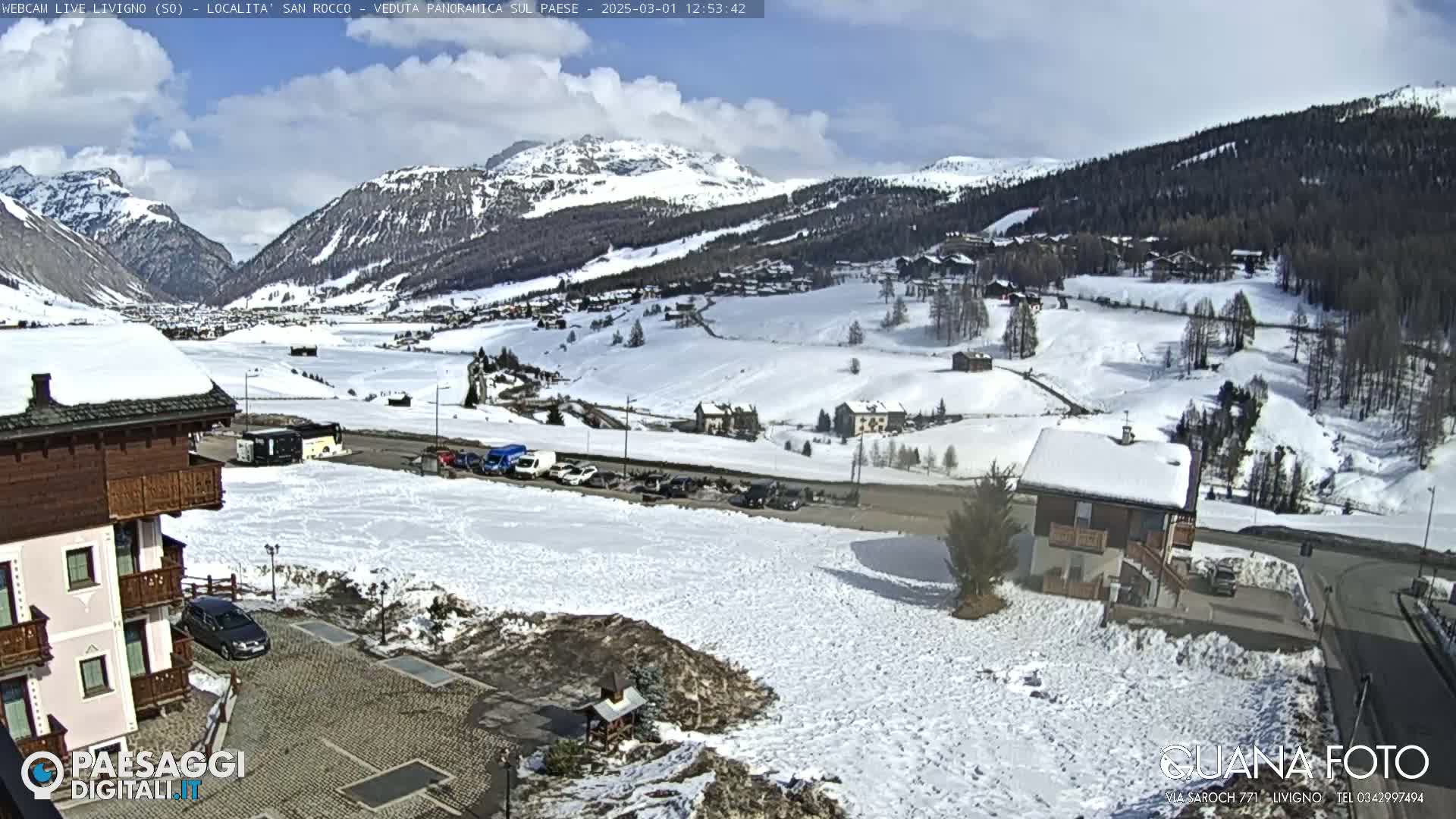 A snow-covered mountain valley village is seen under a partly sunny sky.