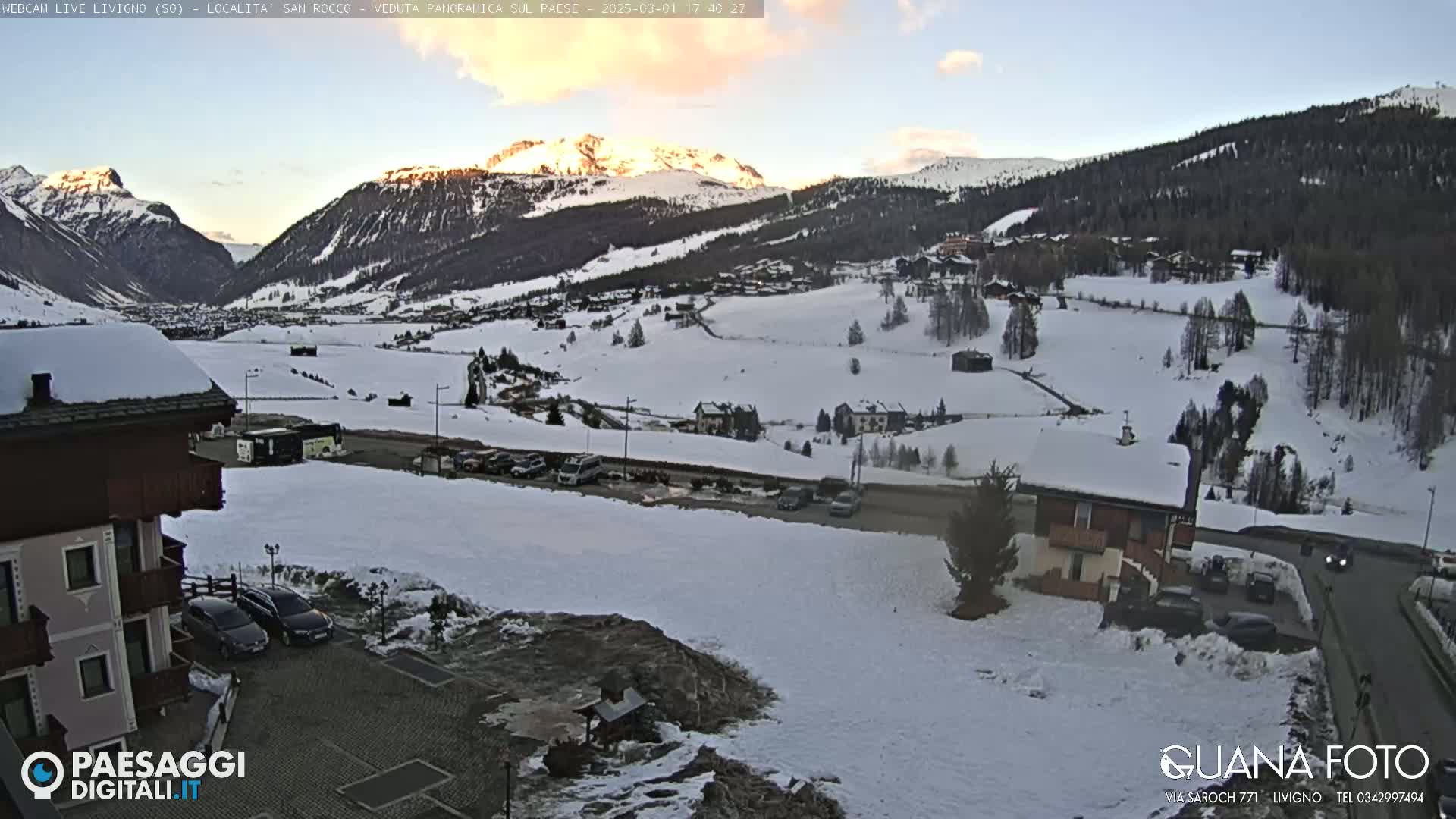 A snow-covered mountain village at sunset, with cars parked in a lot near a building in the foreground.