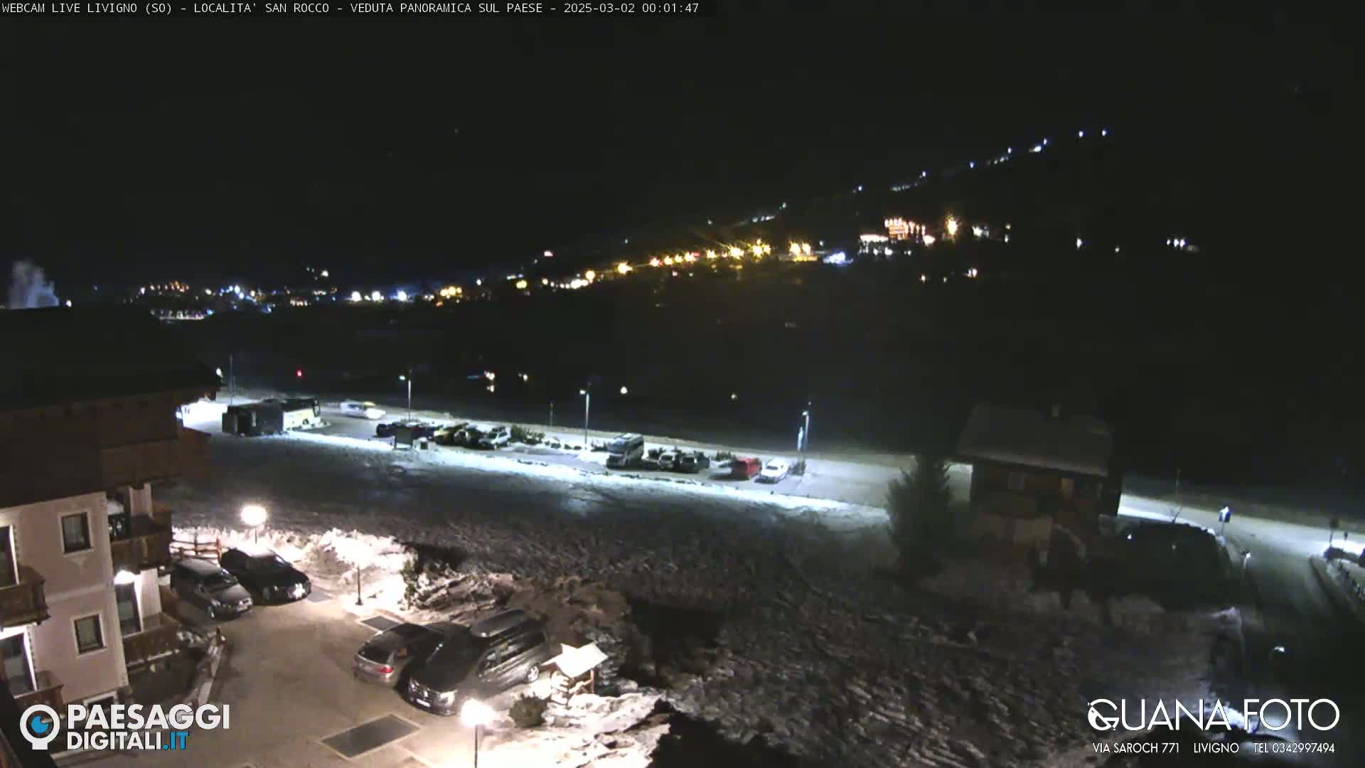 A nighttime, snow-covered parking lot and building are illuminated, with a hillside of lights visible in the distance.