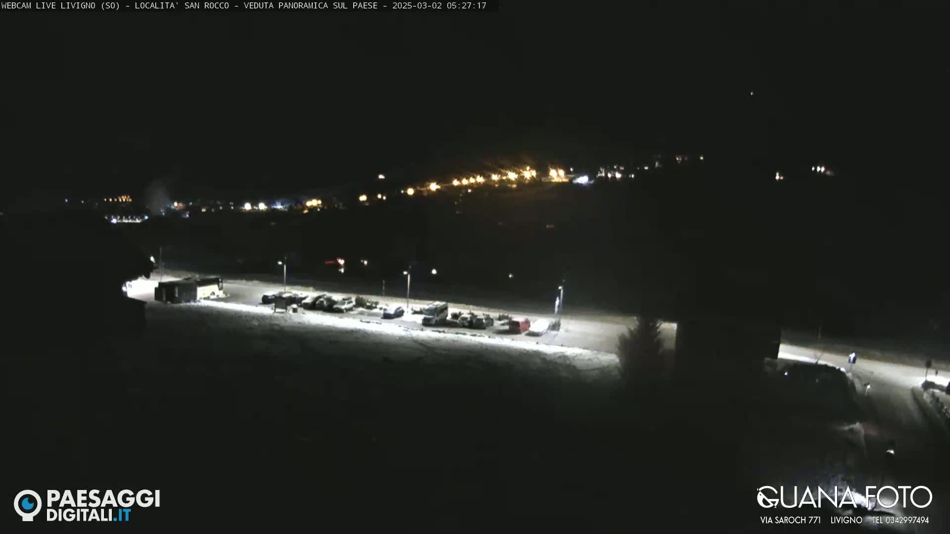 A nighttime, snow-covered parking lot with a few vehicles and distant lights from a town in the background.