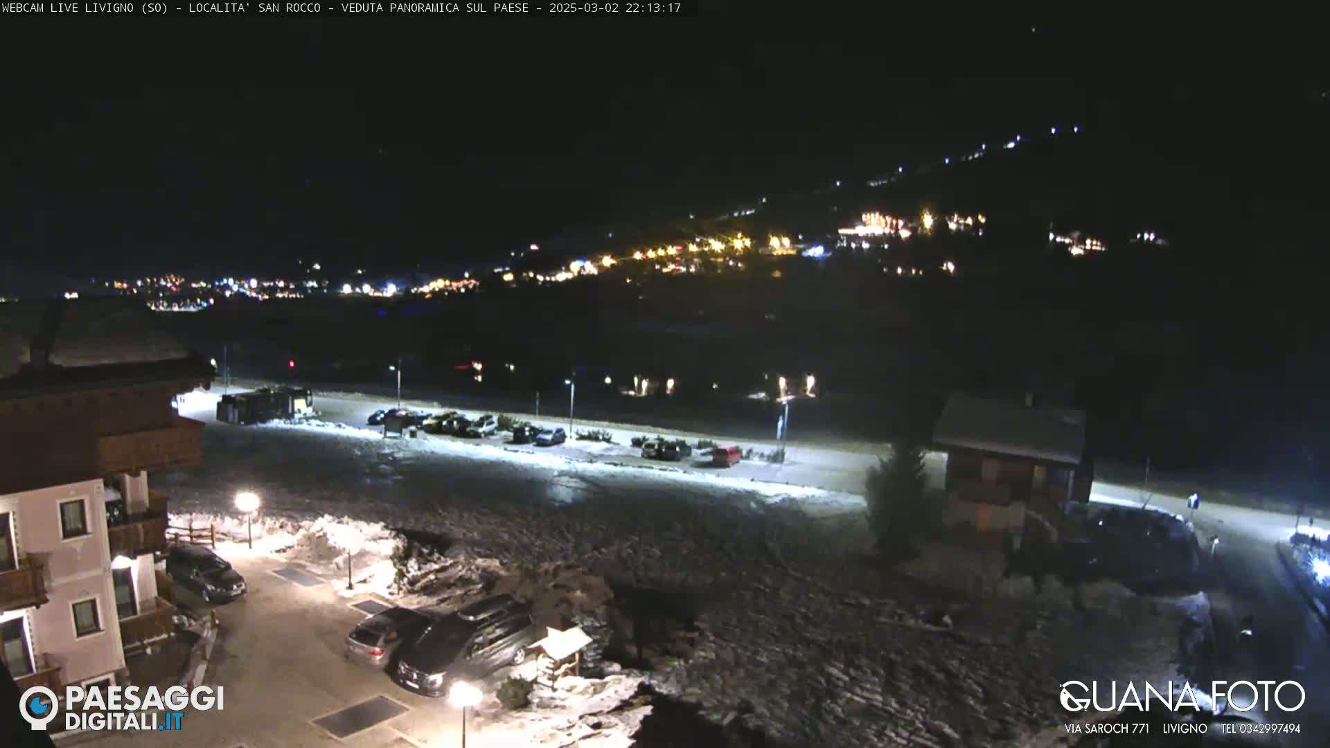 A nighttime, snow-covered parking lot and village are illuminated by lights, with a hillside of lights visible in the distance under a clear, dark sky.