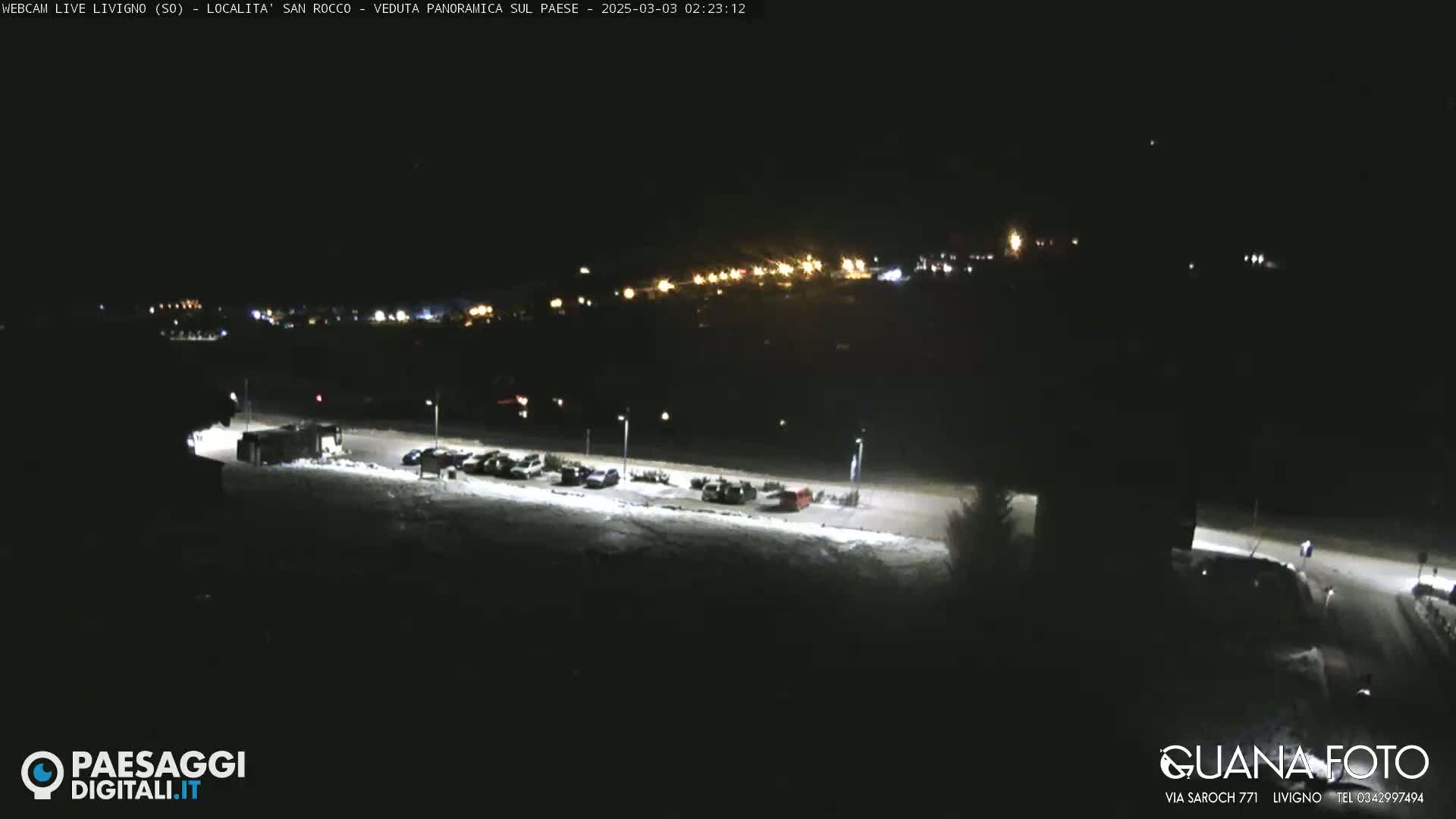 A nighttime, snow-covered parking lot with a few cars is visible, overlooked by a dimly lit town in the distance.