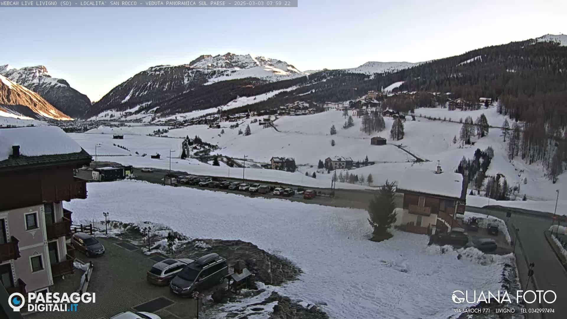 A snow-covered mountain valley town is viewed from a slightly elevated position, with several cars parked near a building in the foreground, under a clear, bright sky.