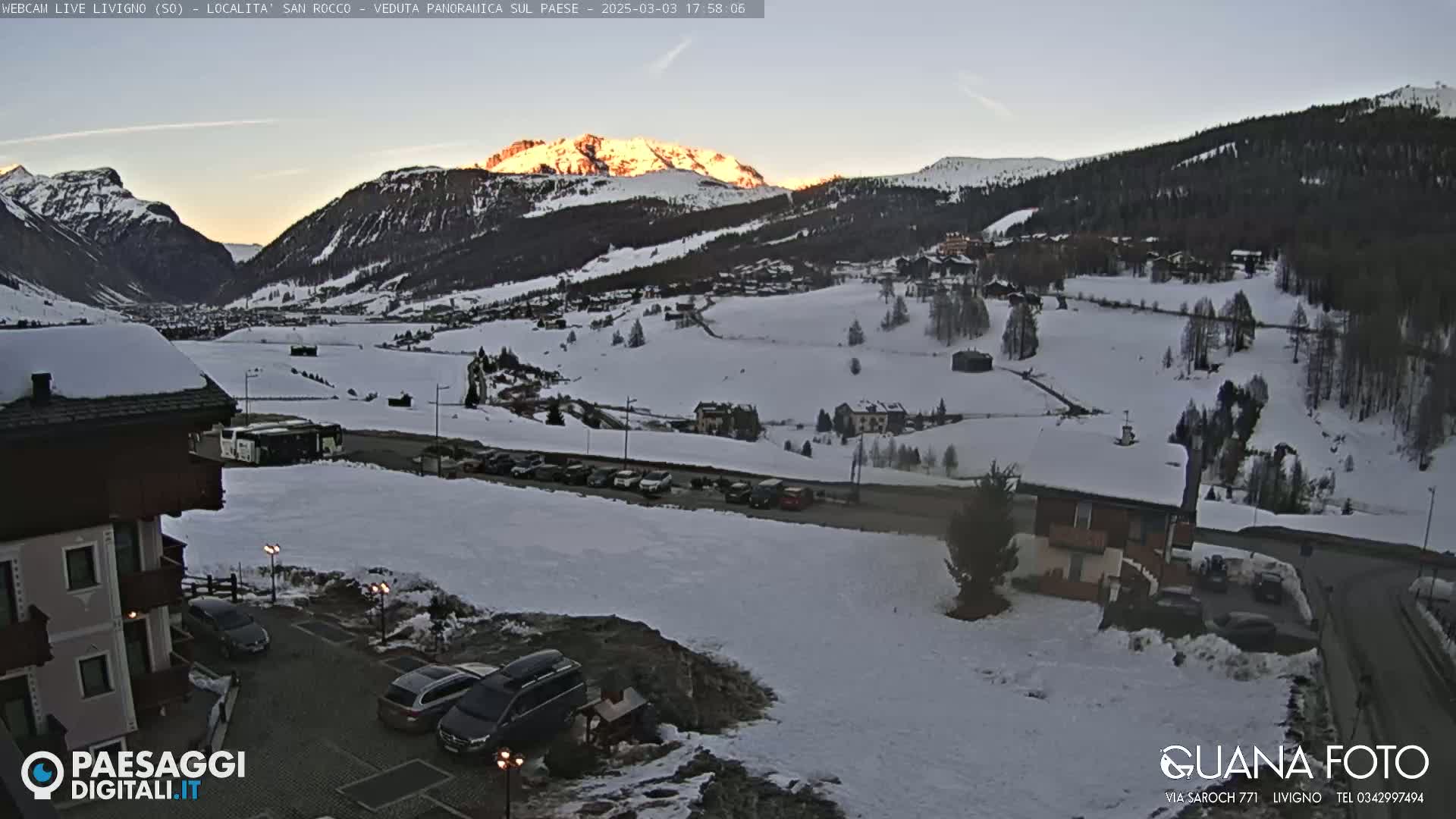 A snow-covered mountain valley at sunset, with cars parked near a building in the foreground.