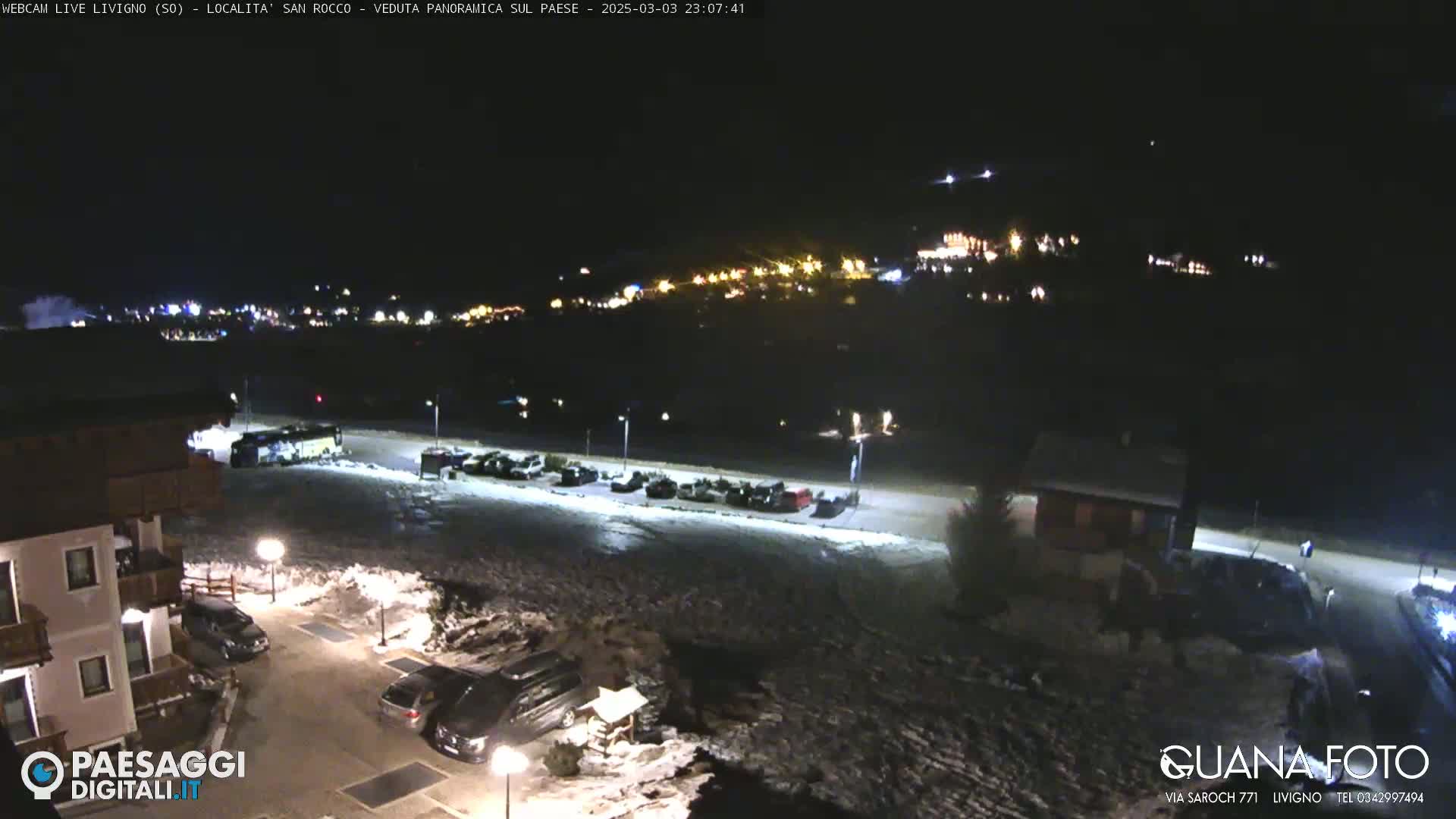 A nighttime, snow-covered parking lot and residential area are shown with a distant, illuminated village in the background.
