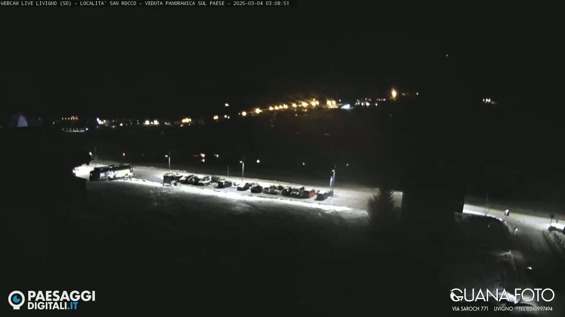 A nighttime, snow-covered parking lot with a few vehicles and a distant, lit town under a clear sky.