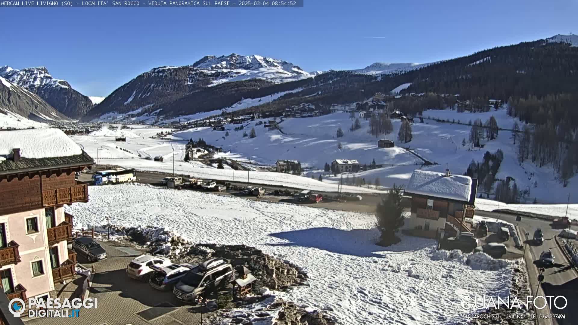 A snow-covered mountain valley town is seen on a sunny day, with cars parked near buildings and snow-covered fields stretching towards the mountains in the background.