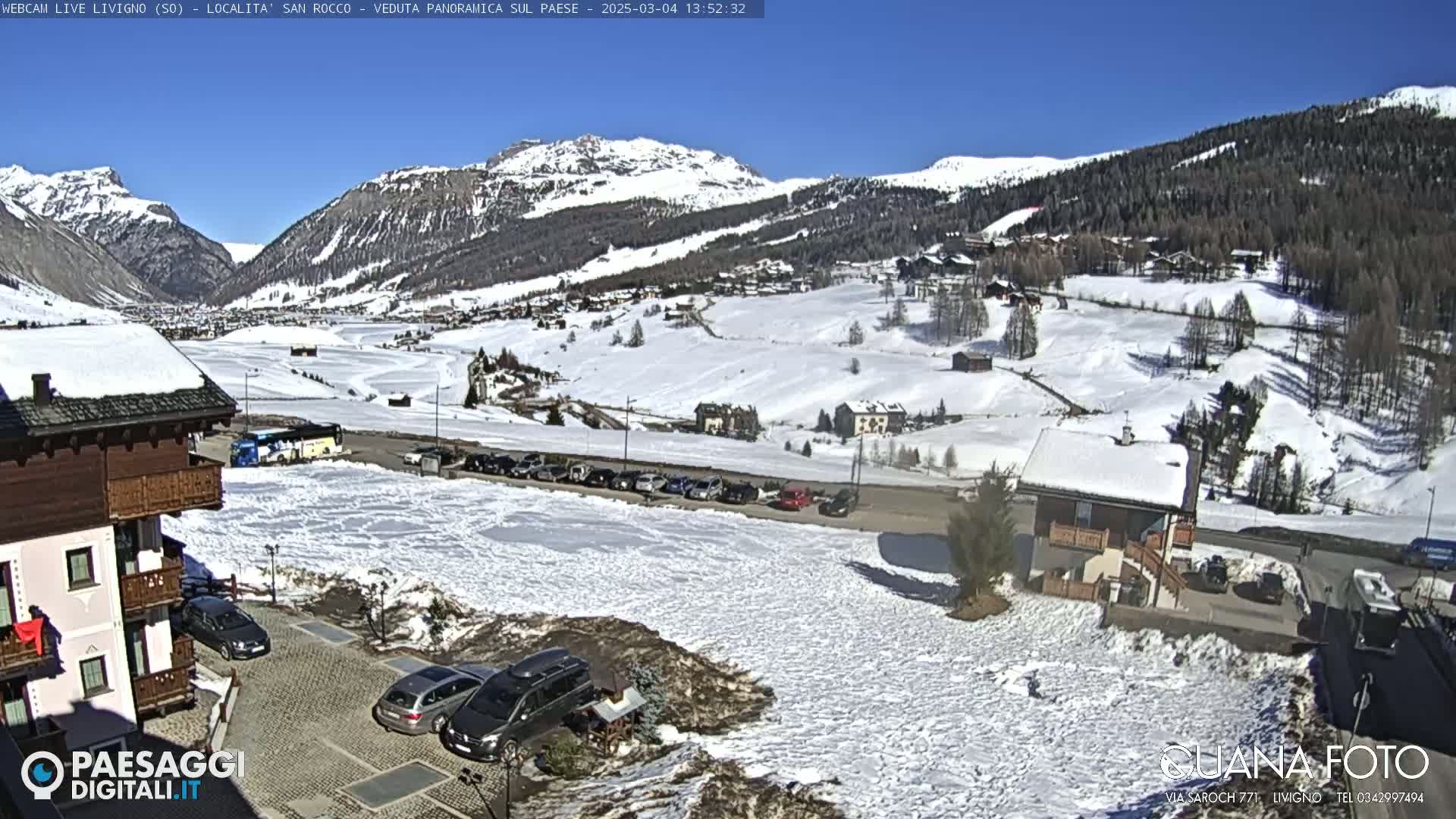 A sunny, snow-covered mountain village scene with several cars parked near buildings, viewed from a slightly elevated position.