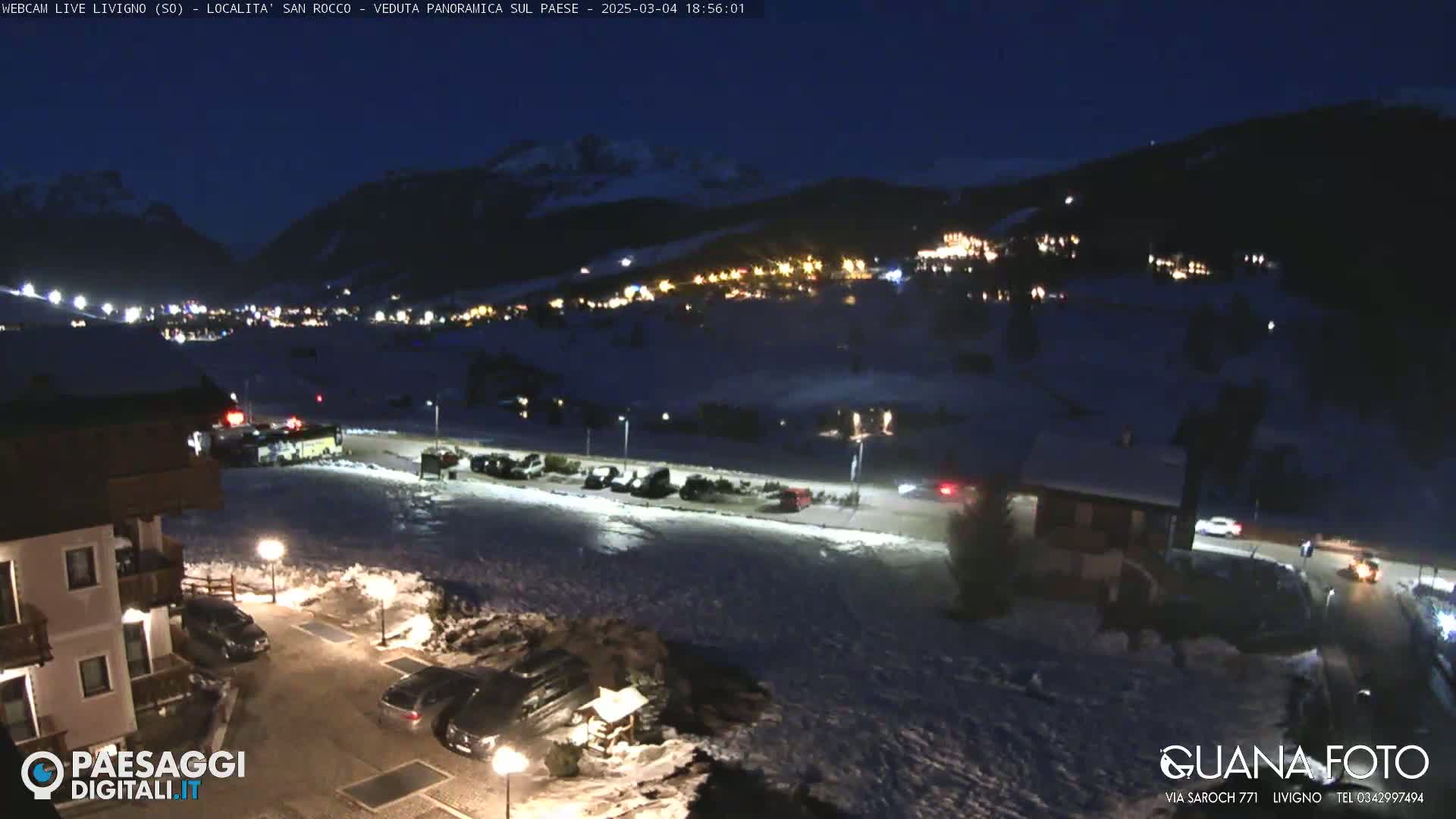 A nighttime view of a snow-covered town nestled in a valley between mountains, with some cars parked in a lot in the foreground.